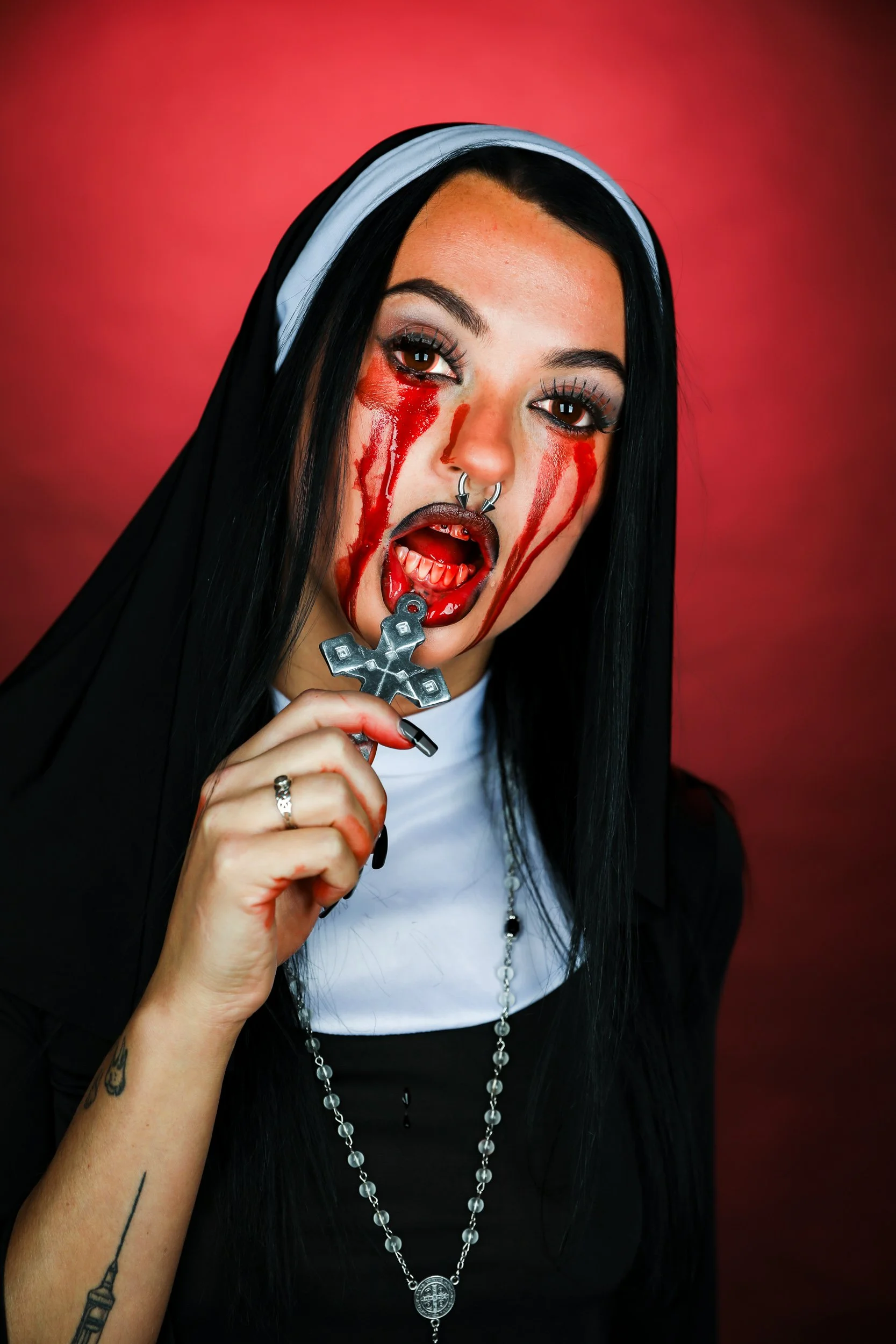 A woman with long black hair and religious tattoos holding a silver cross necklace, with blood streaming down her face and mouth, dressed in a nun's habit against a red background.