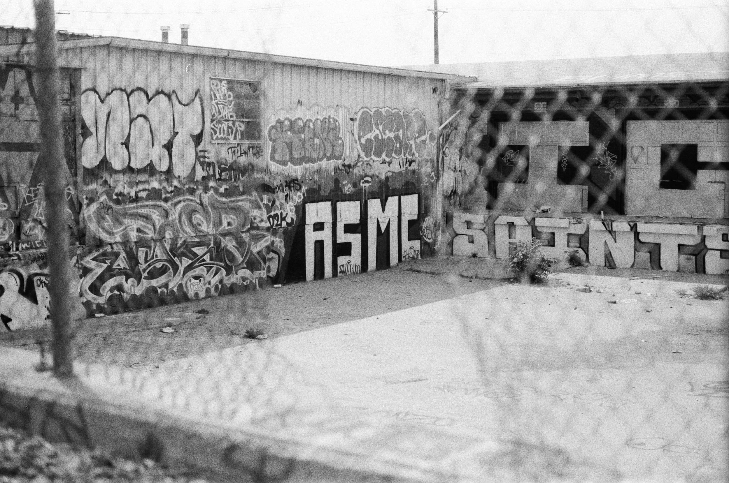 A black and white photo of a graffiti-covered building behind a chain-link fence. The building appears abandoned, with boarded-up windows and large painted letters on the wall.