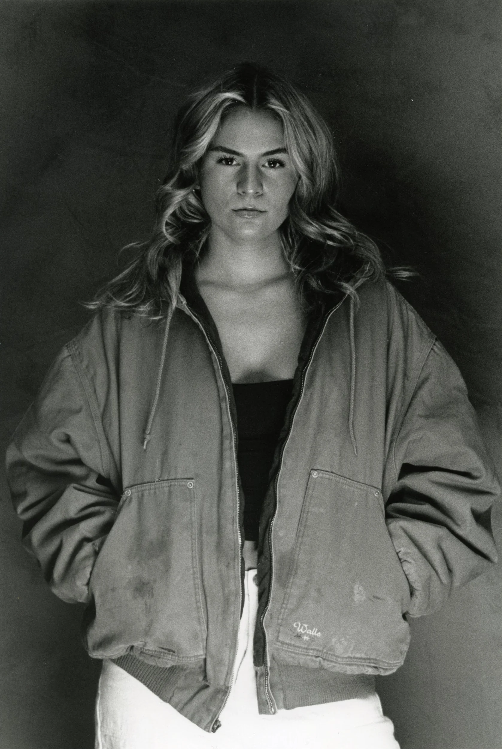 A young woman with wavy hair wearing a casual jacket and white pants, standing against a dark background.
