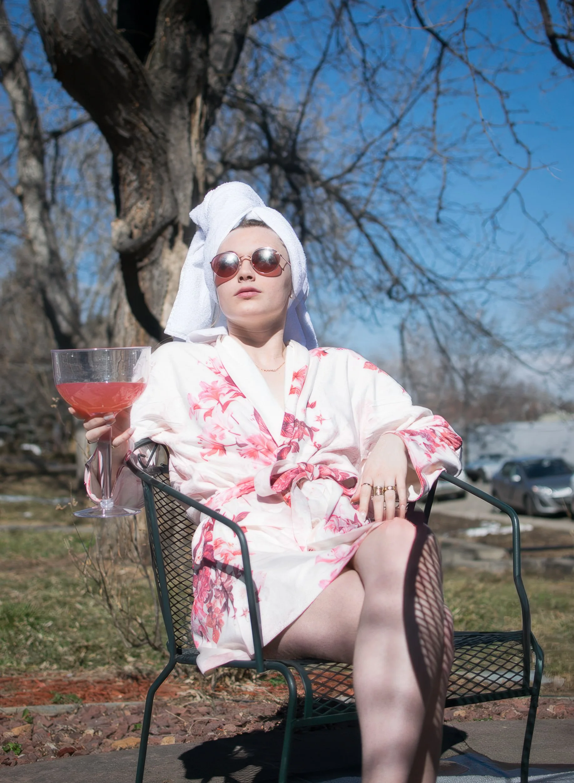 A woman wearing sunglasses, a towel on her head, and a floral robe, sitting outdoors on a metal chair, holding a large glass of pink beverage, with a tree and parked cars in the background on a sunny day.