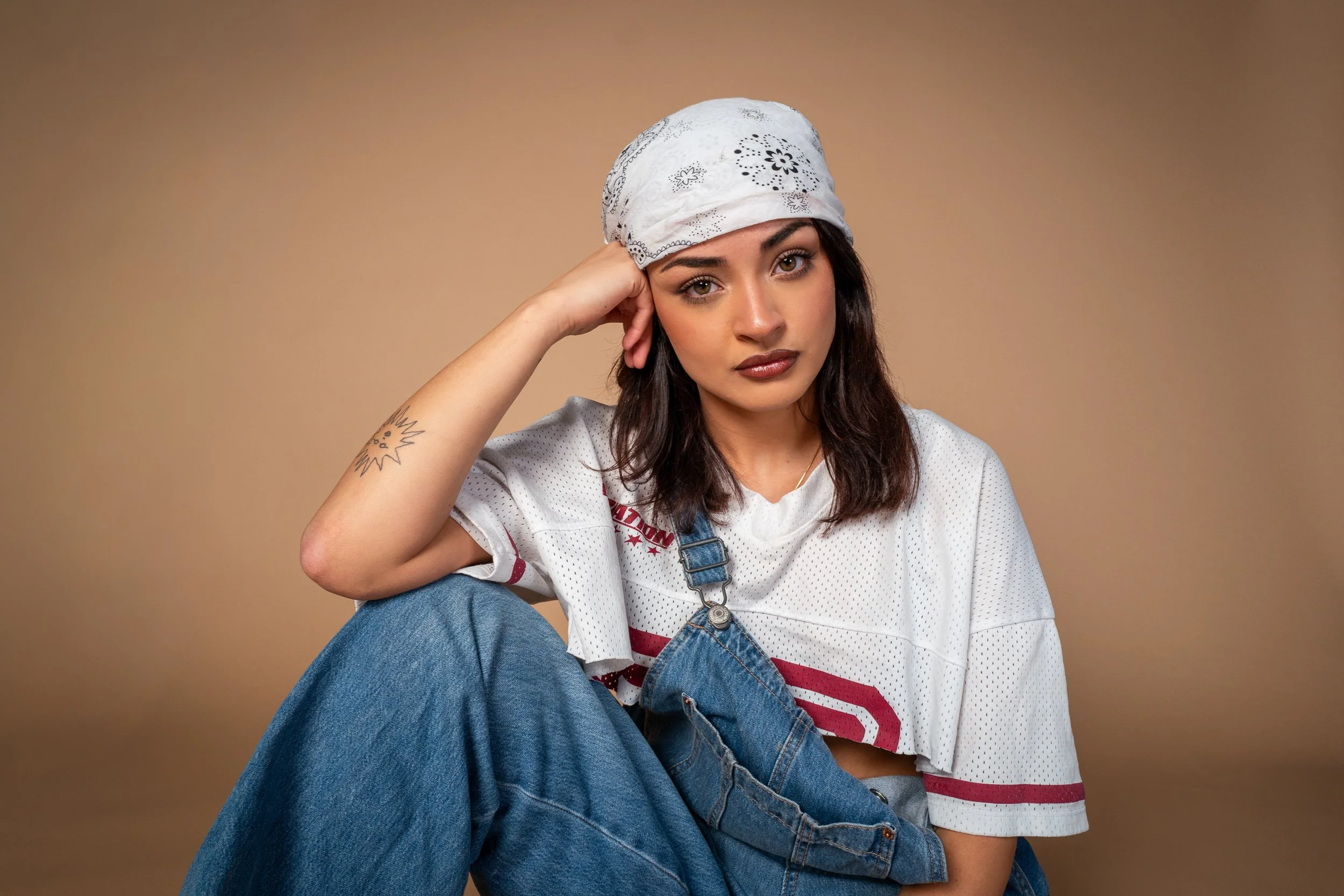 A young woman with dark hair and light skin, wearing a white bandana with black patterns, a white sports jersey with red accents, and blue jeans, sitting against a plain beige background. She has her head resting on her left hand and looks directly at the camera with a neutral expression.
