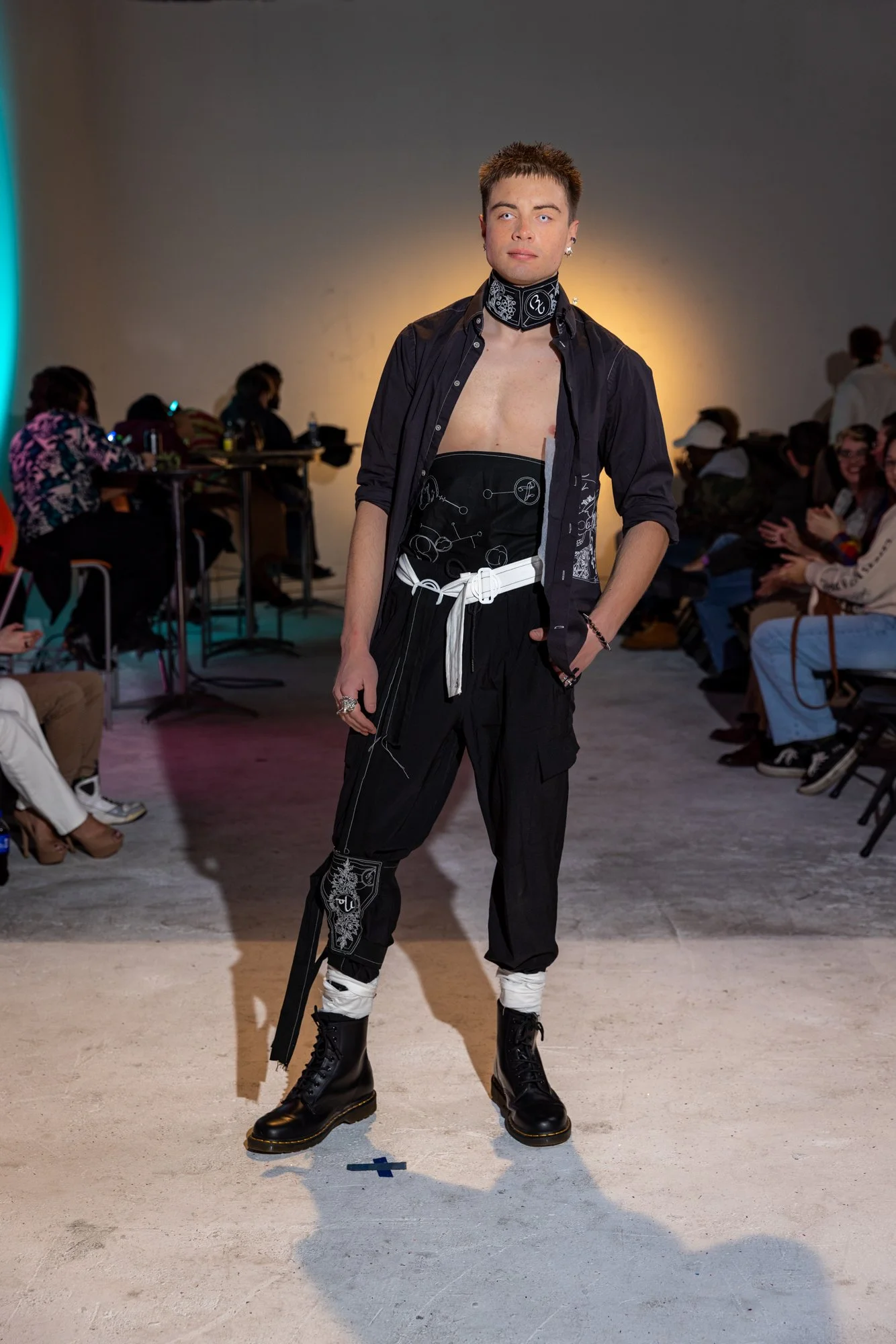 A male model walking on a runway during a fashion show, wearing black pants, a black patterned shirt, a black bandana with white designs, and black combat boots. The background shows an audience seated on both sides of the runway.