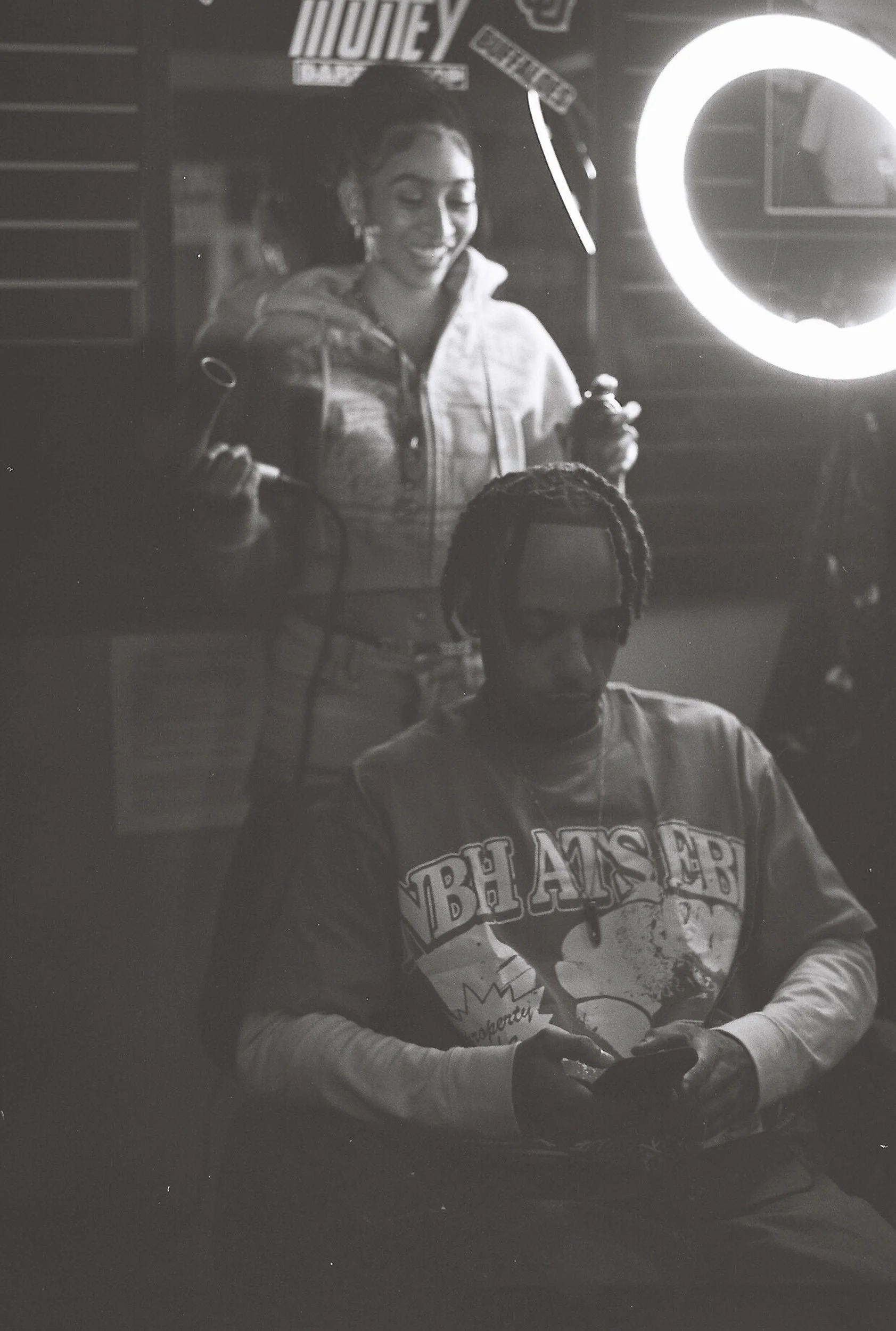 A woman with long hair tied back, smiling and holding a golf club, is standing next to a ring light. She is wearing a hoodie and appears to be in a recording or streaming setup. A person with dreadlocks, wearing a sweatshirt with a comic style printe