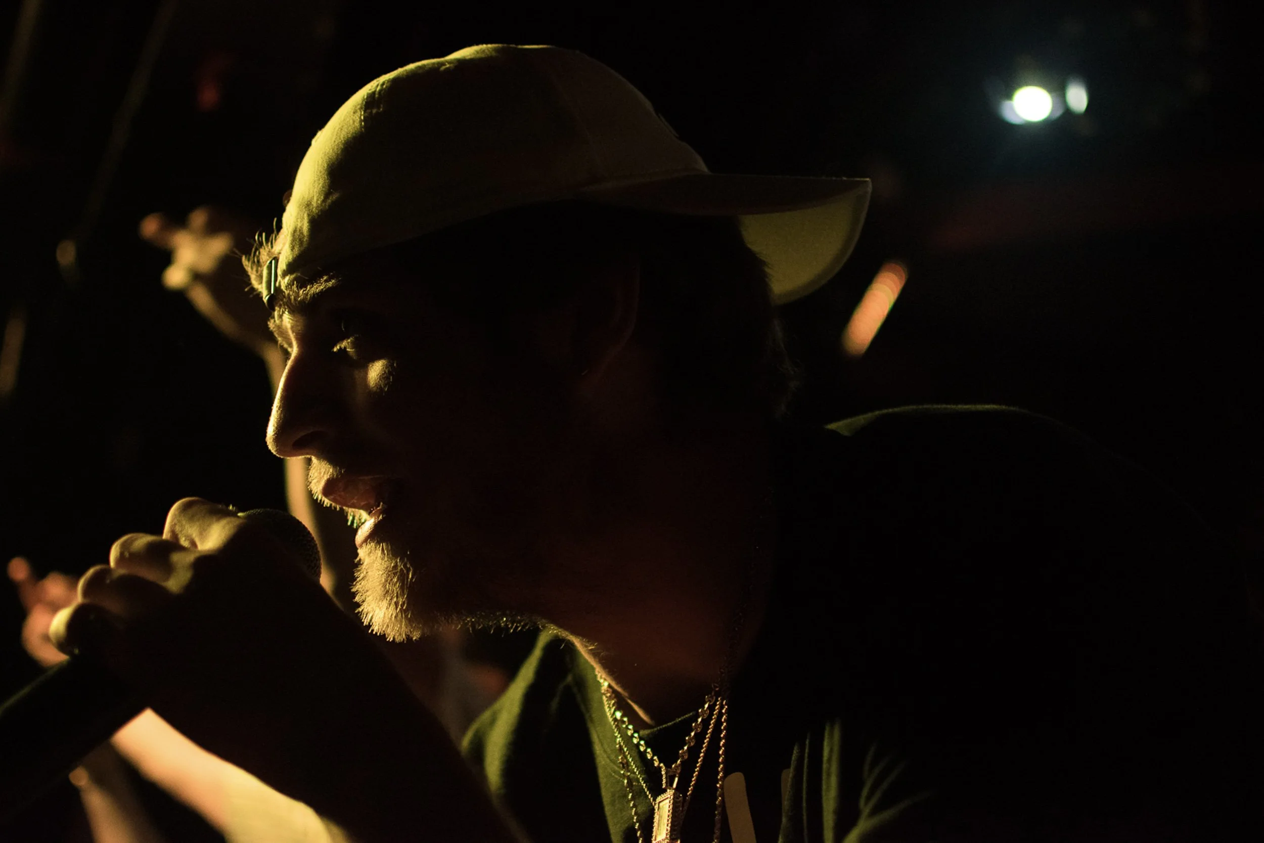 A person wearing a cap, illuminated by warm light, with visible jewelry around their neck, against a dark background with some bright light spots.