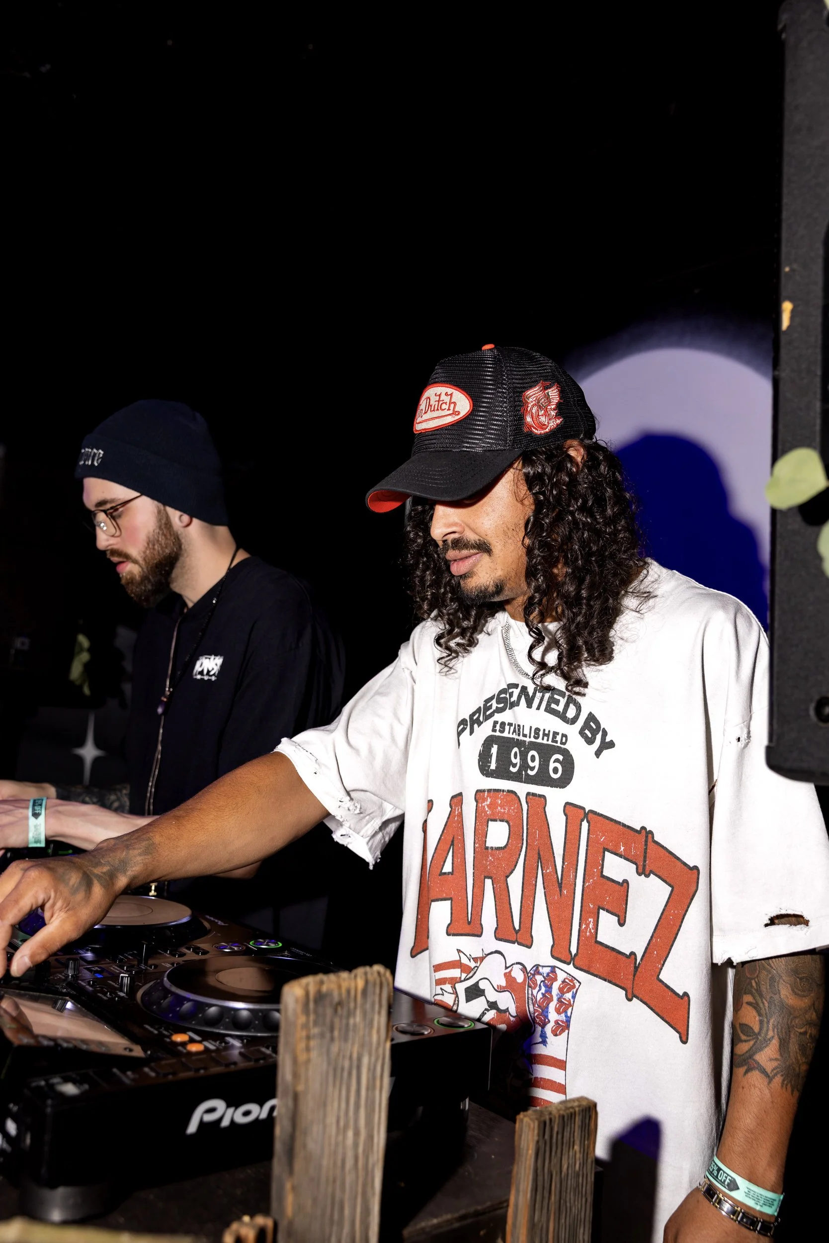 Two men are DJing at an event indoors. The man on the right has curly hair, a mustache, and a goatee, wearing a black and red baseball cap with a logo, a white T-shirt with graphic and text, and tattoos on his arms. The man on the left has glasses, a