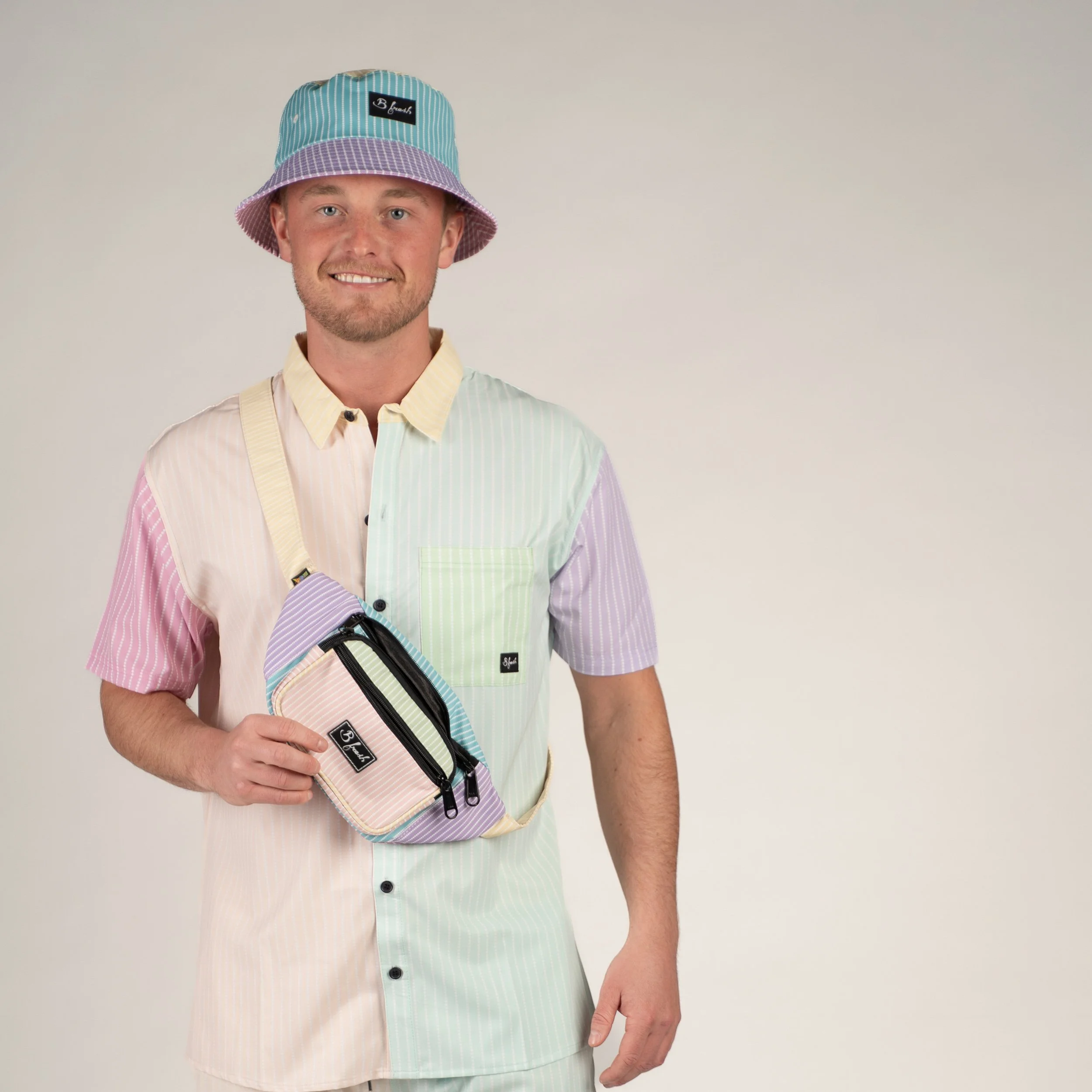 A young man wearing a pastel-colored, striped shirt and a multicolored bucket hat, holding a small bag.