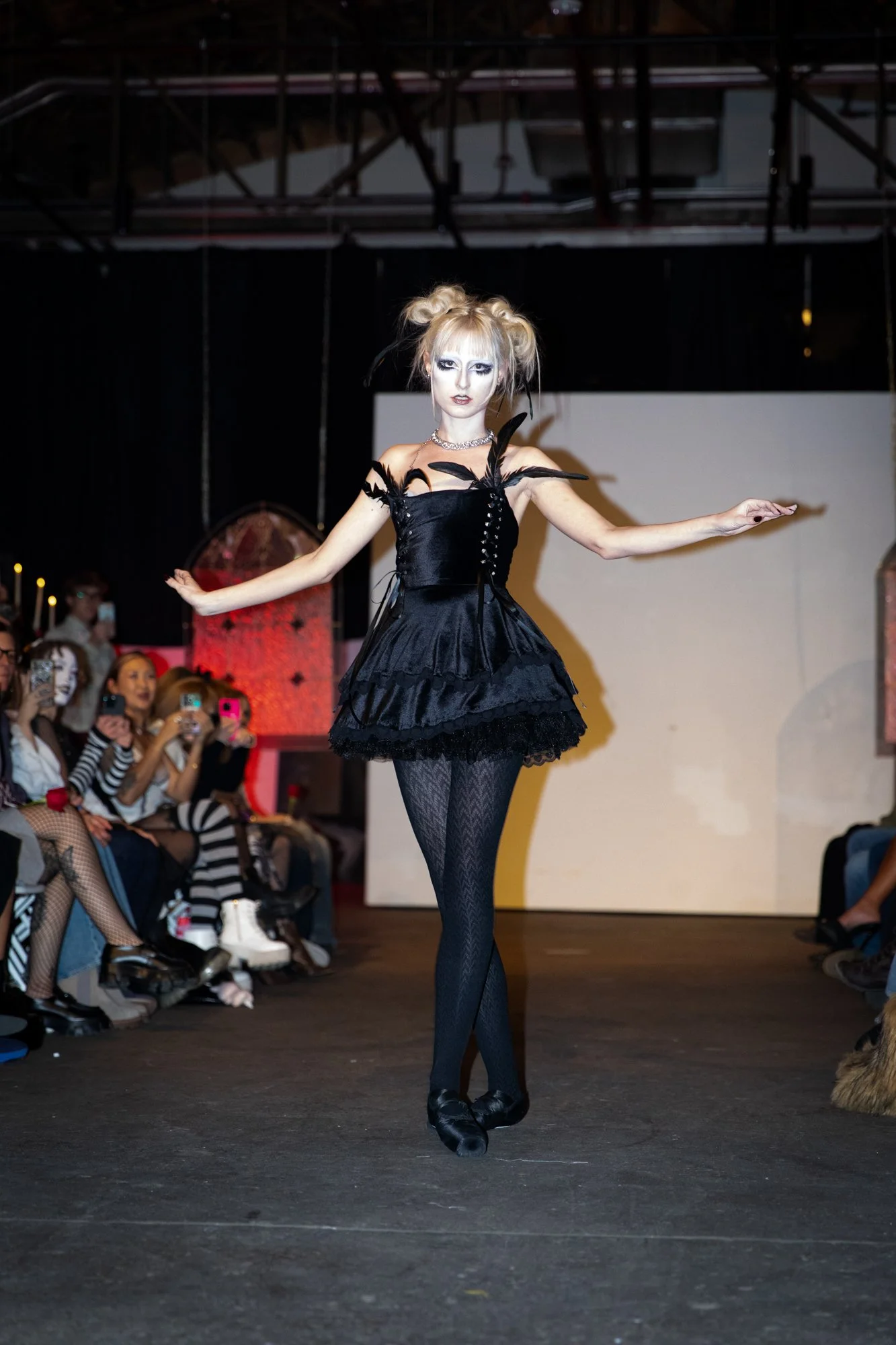 A model is walking on a runway wearing a black gothic-inspired dress with feathers, lace stockings, and black shoes. The audience watches and takes photos.