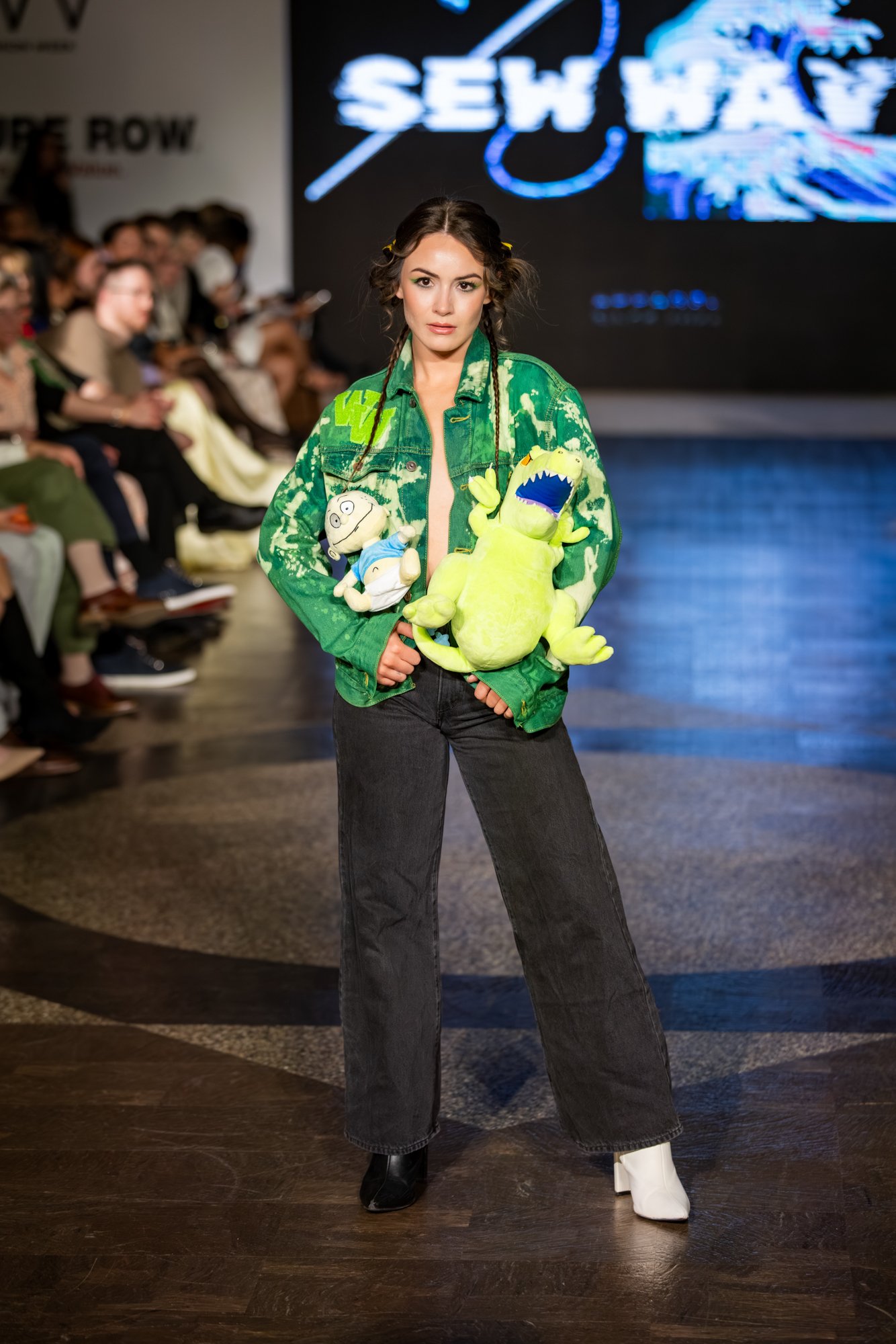 A woman on a runway wearing a green tie-dye jacket and black jeans, holding plush toys, with an audience and a large screen in the background.