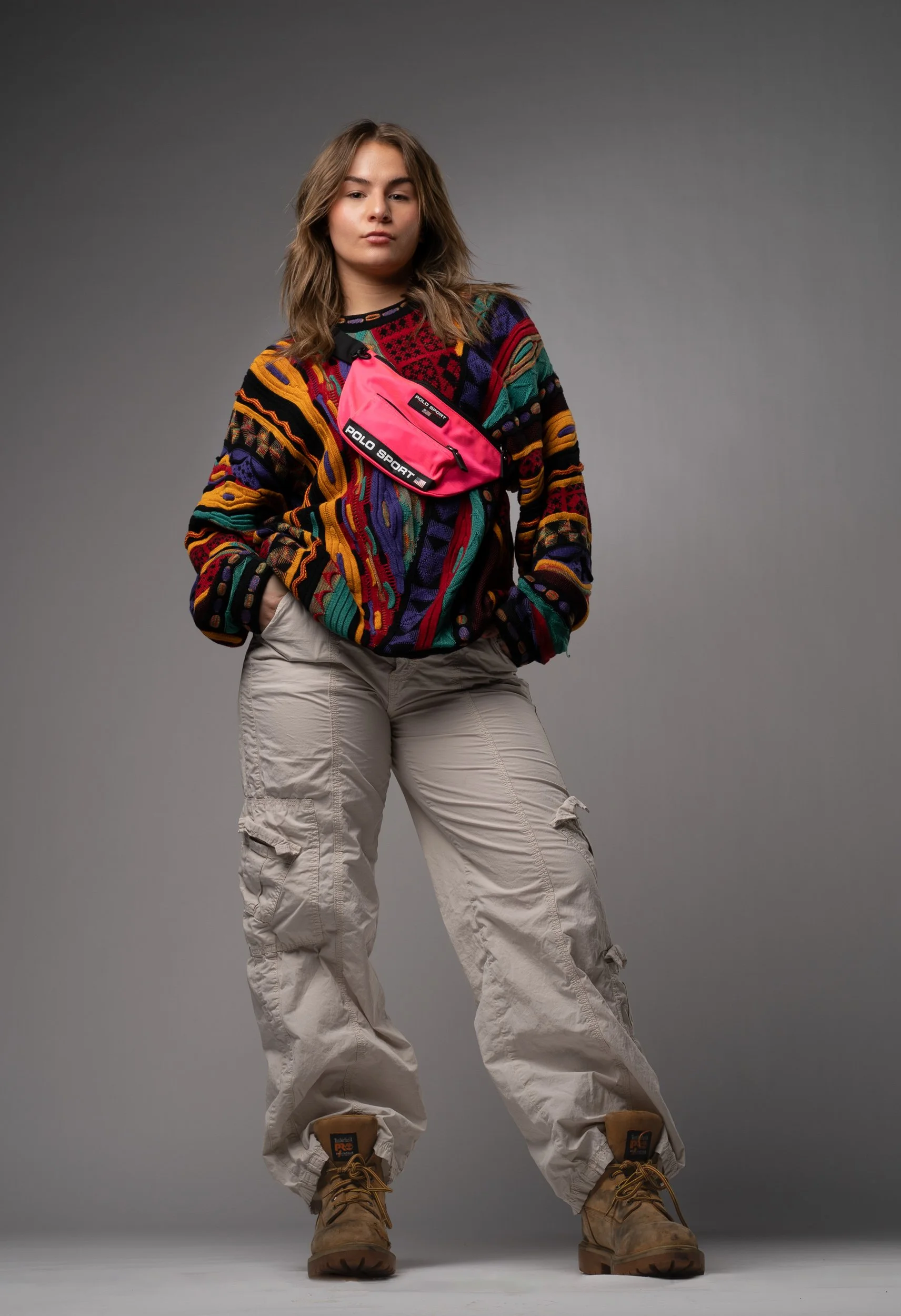 A young woman with shoulder-length wavy hair poses confidently against a plain gray background. She is wearing a colorful, patterned sweater, beige cargo pants, and tan work boots. She has a pink crossbody bag with black and white labels reading "POL
