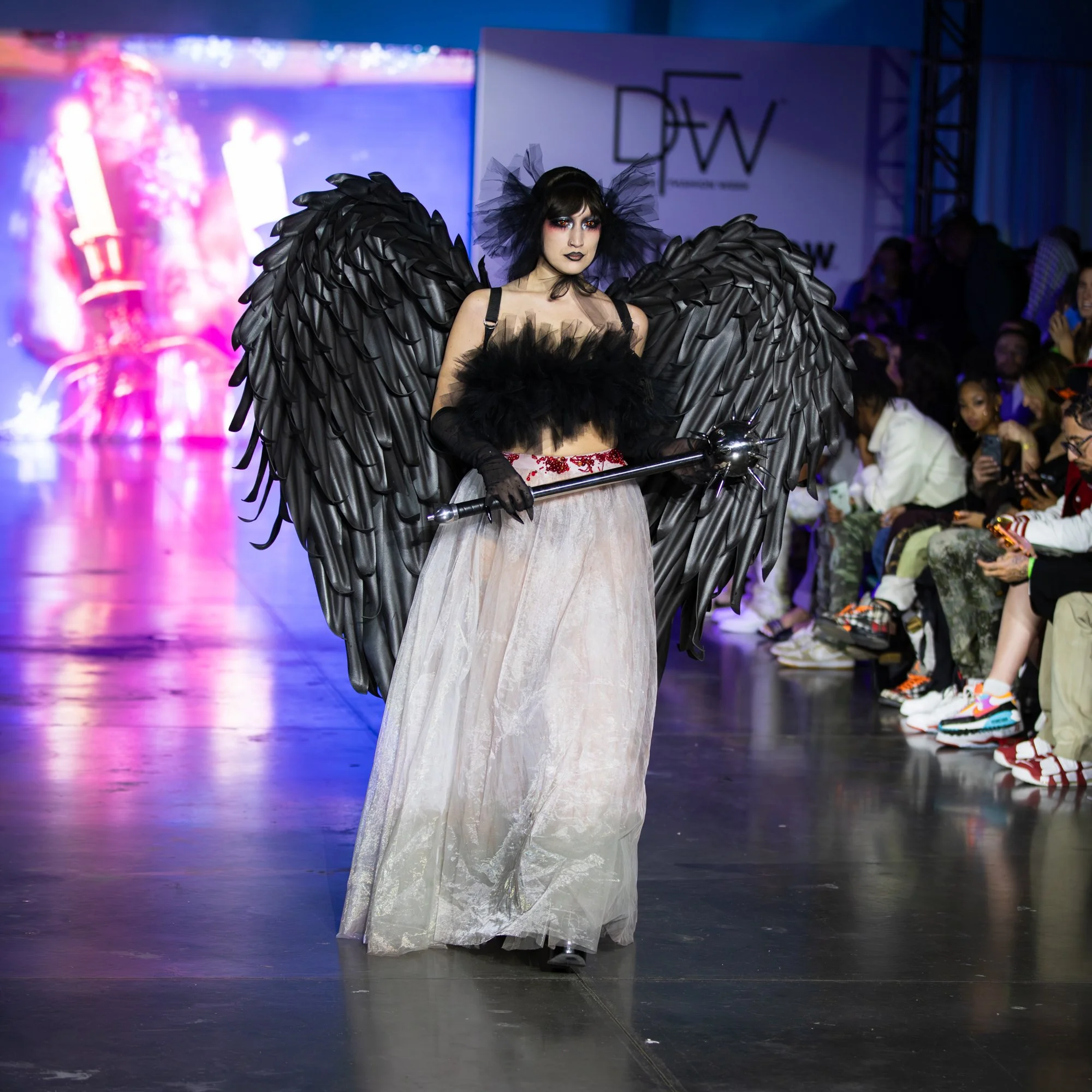 Person on a runway wearing a black feathered angel costume with large wings, a tulle headpiece, black makeup, and a staff, at a fashion show.