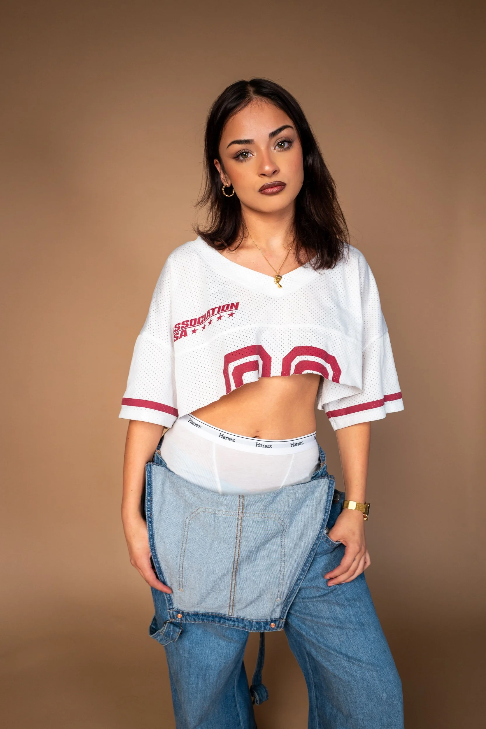 Young woman with light to medium skin tone and shoulder-length dark hair, wearing a white sports jersey with red lettering and numbers, a pair of high-waisted blue jeans, gold hoop earrings, a gold necklace, and a gold wristwatch, standing against a 