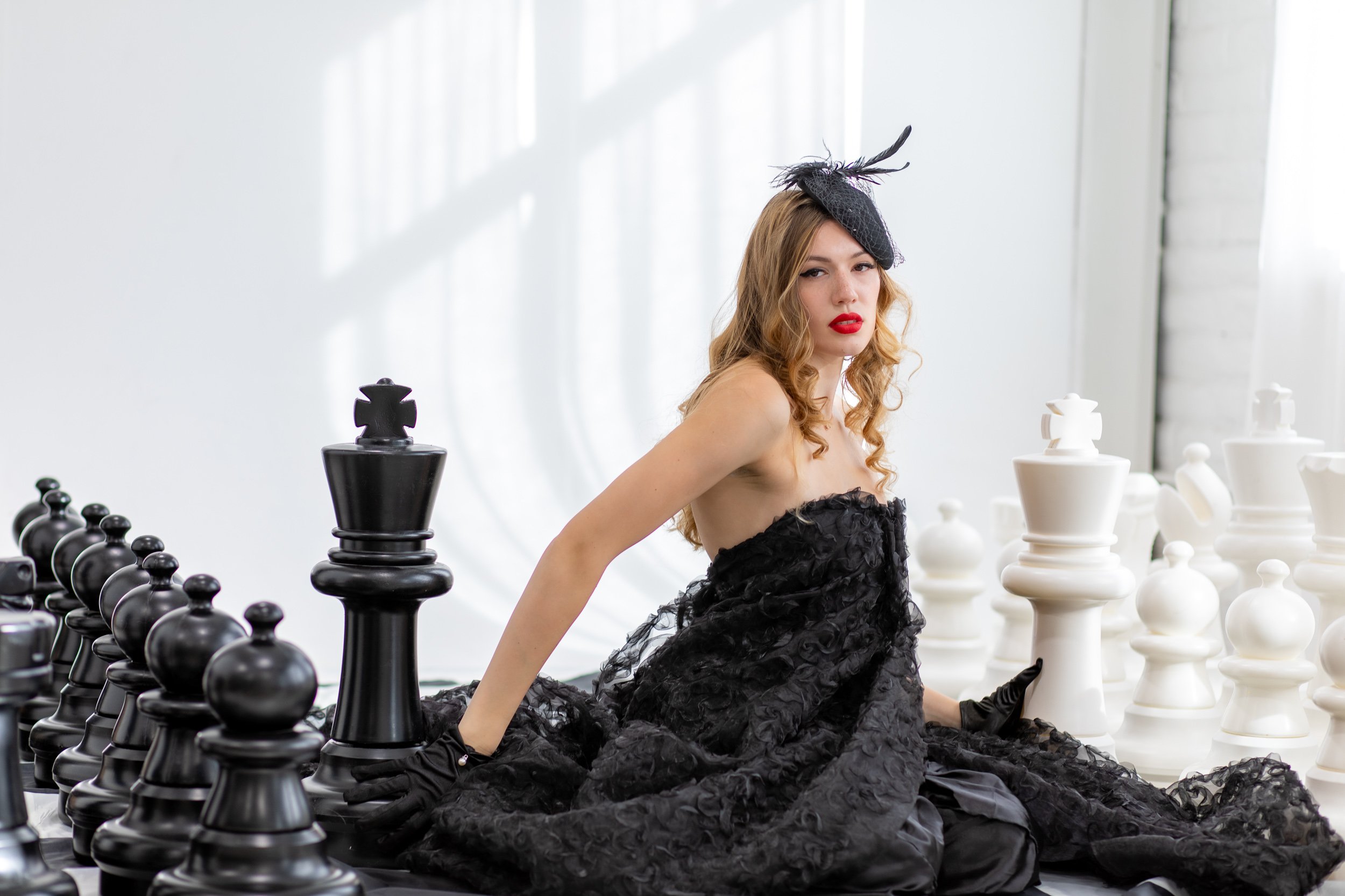 A woman with long, curly hair and red lipstick, wearing a black feathered dress and hat, sitting among large chess pieces, with white and black chess pieces on the table.