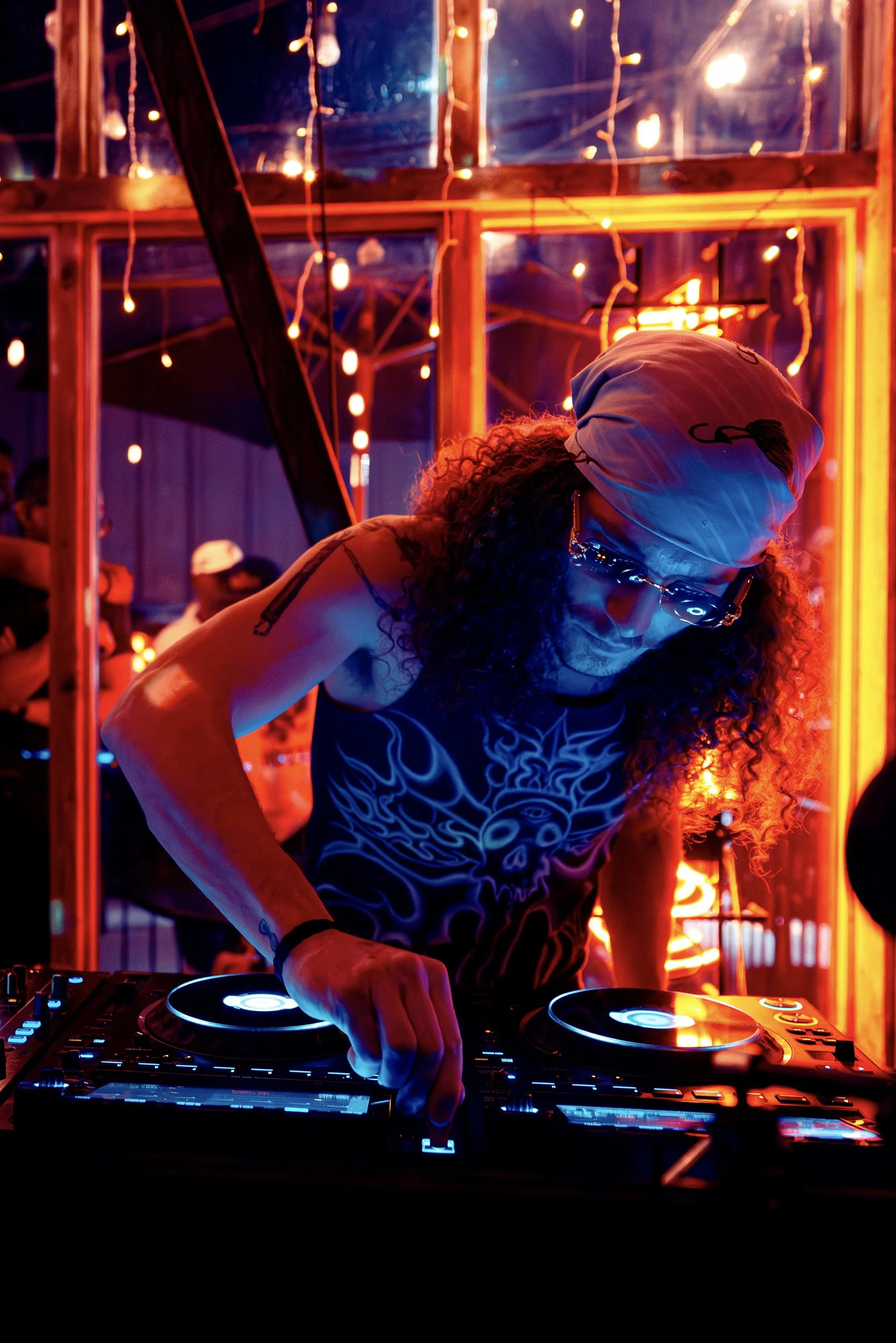 A DJ with curly hair, wearing sunglasses and a beanie, performs at a neon-lit event. The background is illuminated with red and orange lights, and there are people in the background.