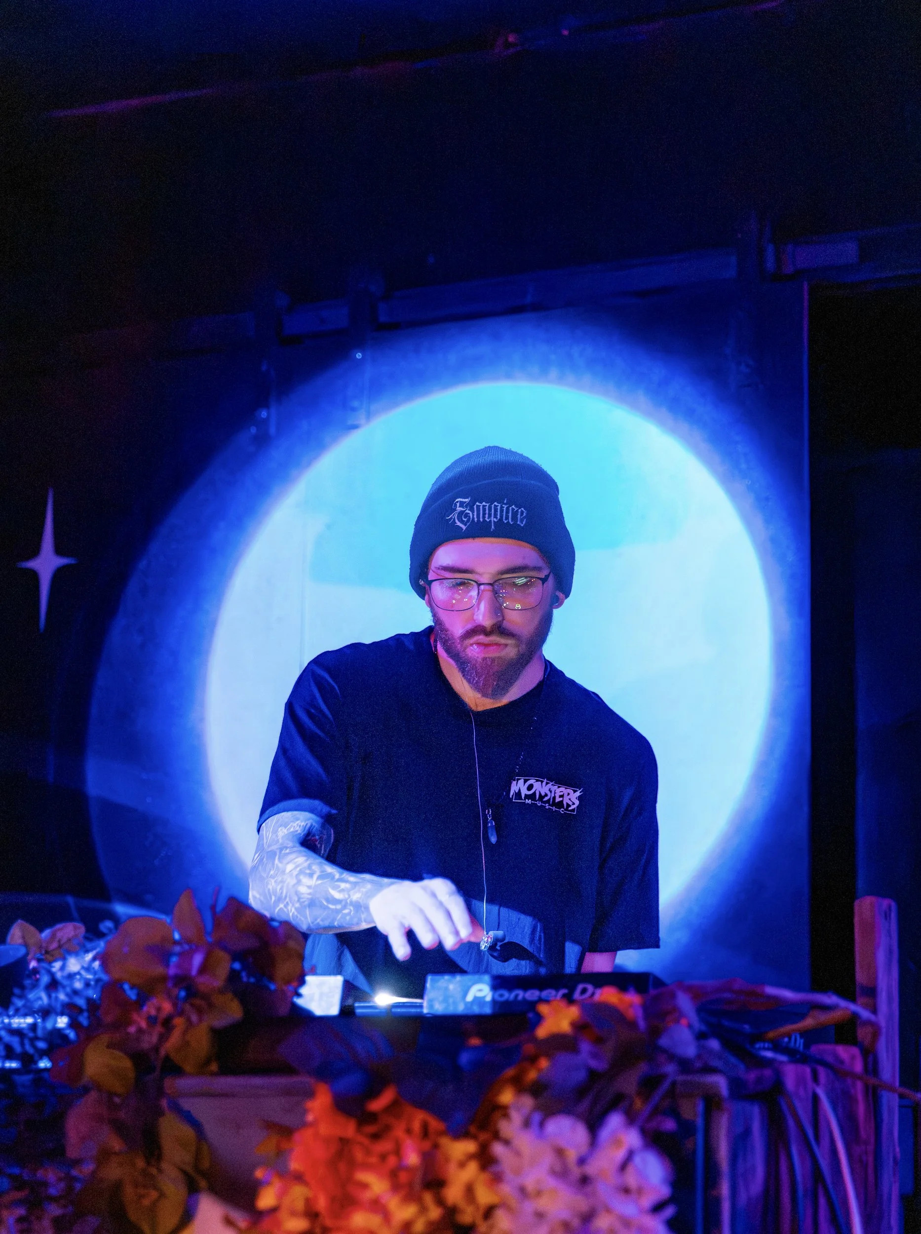 A male DJ with glasses, tattoos, and a beanie reading 'Emoice' performs at a nightclub with blue lighting, surrounded by flowers and a Pioneer DJ setup.