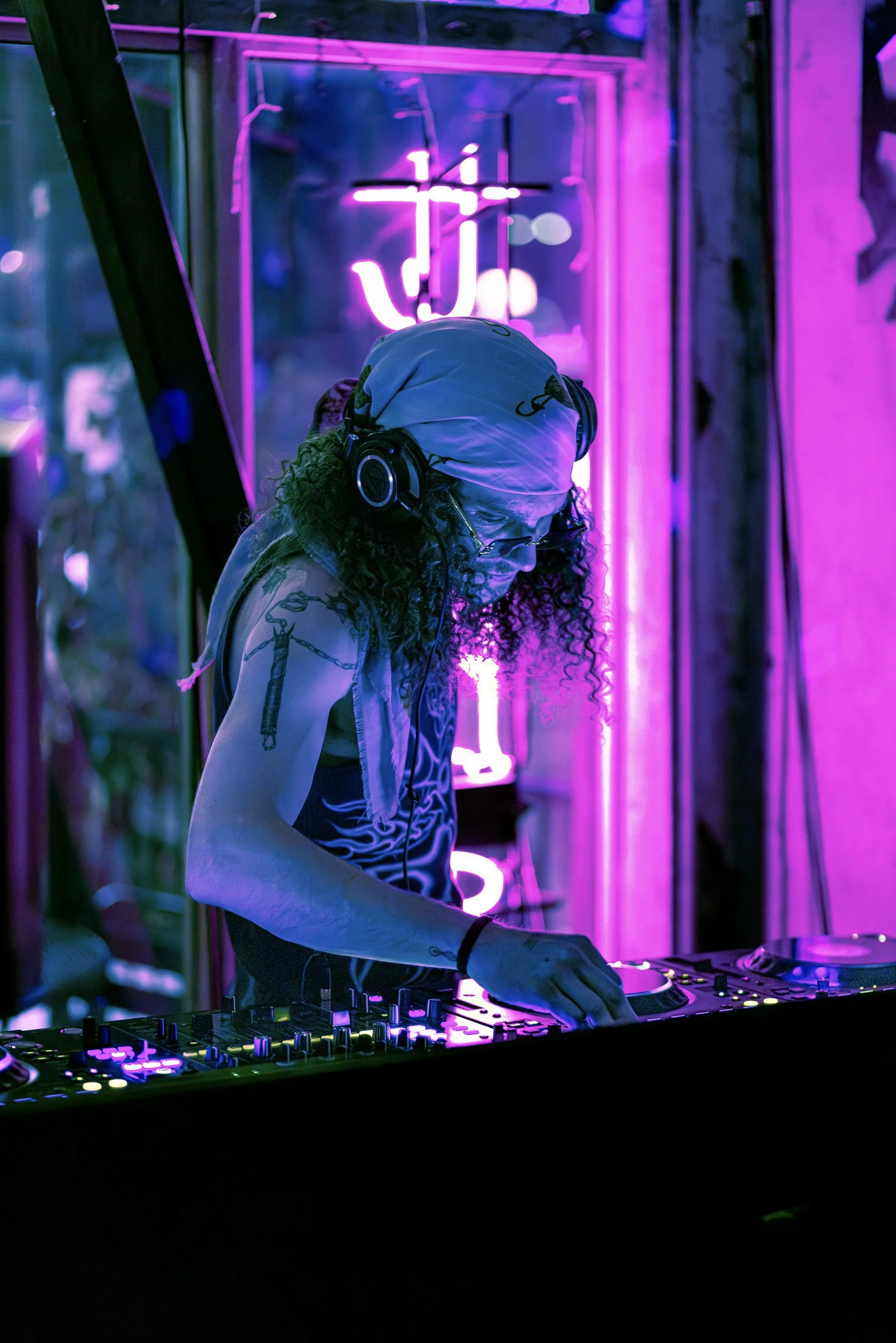 Person with curly hair wearing headphones and a face covering, DJ-ing at a neon-lit venue with purple and pink lighting, in front of a neon cross and lettering.
