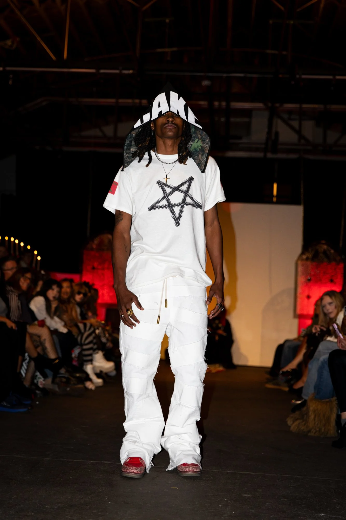 A male model walking down a runway during a fashion show, wearing a white graphic t-shirt with a pentagram design, white pants, and red sneakers. The model's face is partially obscured by a hood with large white letters.