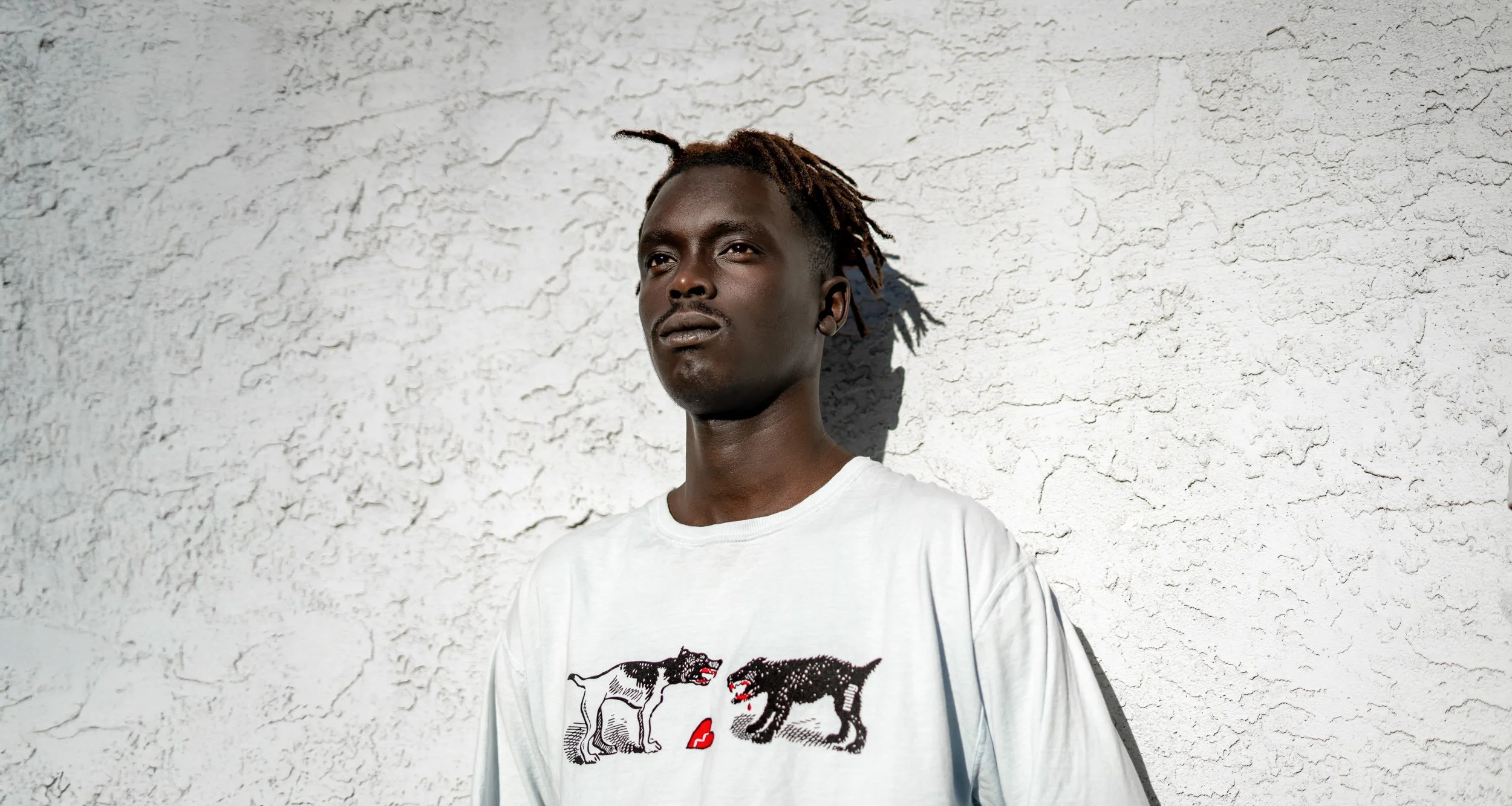 A young man with dreadlocks wearing a white T-shirt with a graphic of two dogs facing each other standing against a textured white wall.