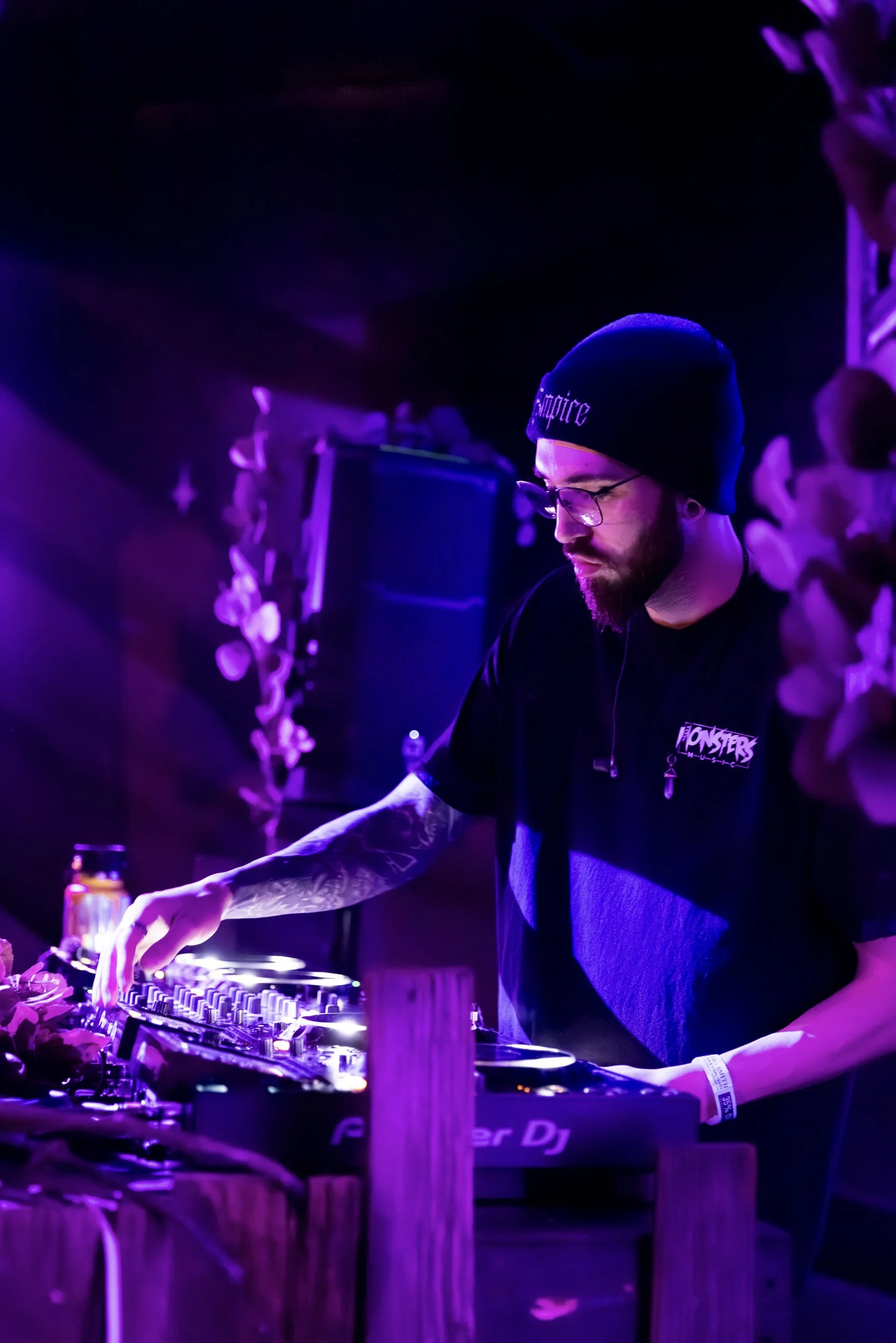 A DJ performing at a nightclub with purple lighting, wearing glasses, a beanie, and a black t-shirt that says 'Monsters'.