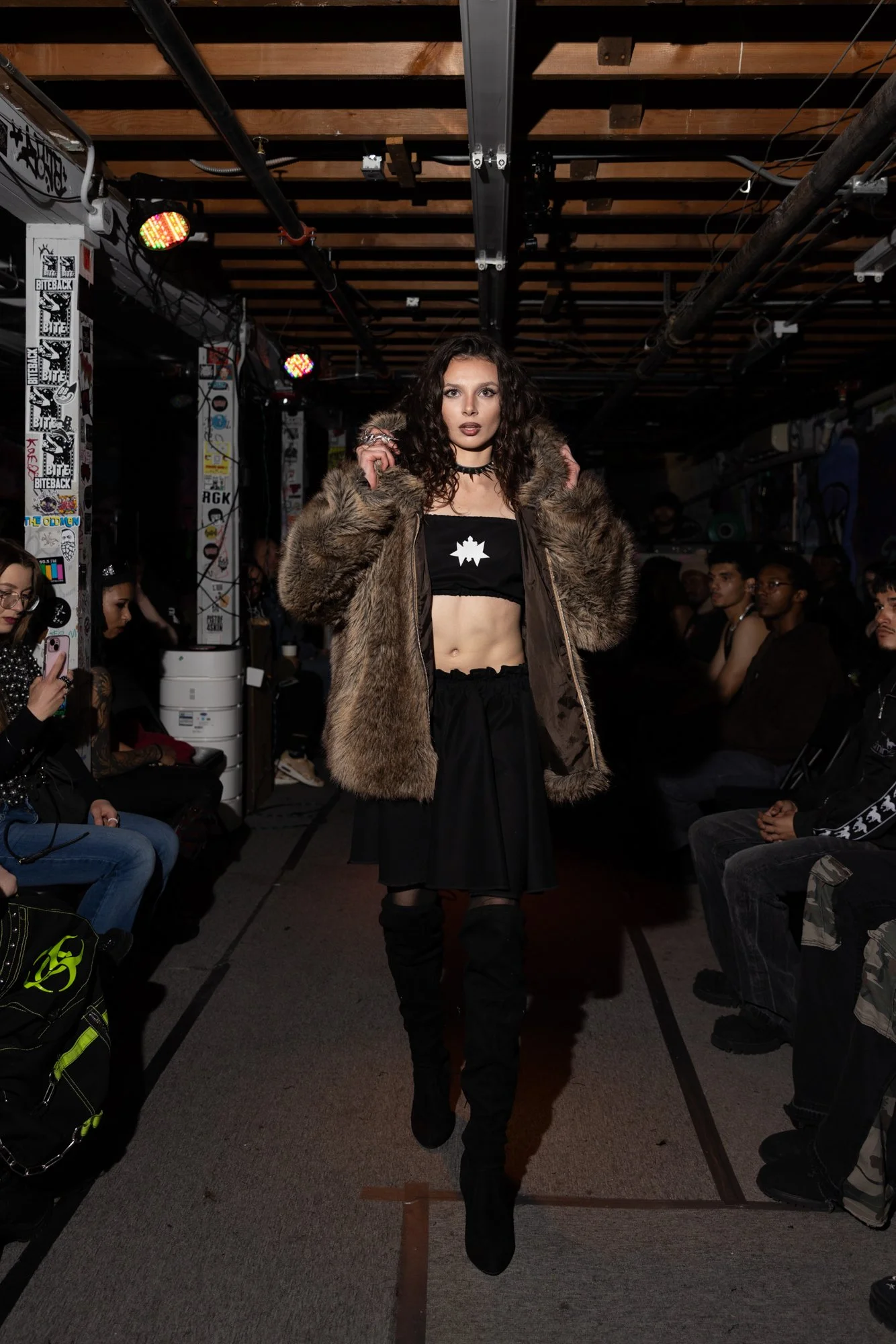 A woman with curly hair and dark makeup walking down a runway during a fashion show. She is wearing a fur-lined jacket, a crop top, and black pants, with an audience seated on both sides of the runway