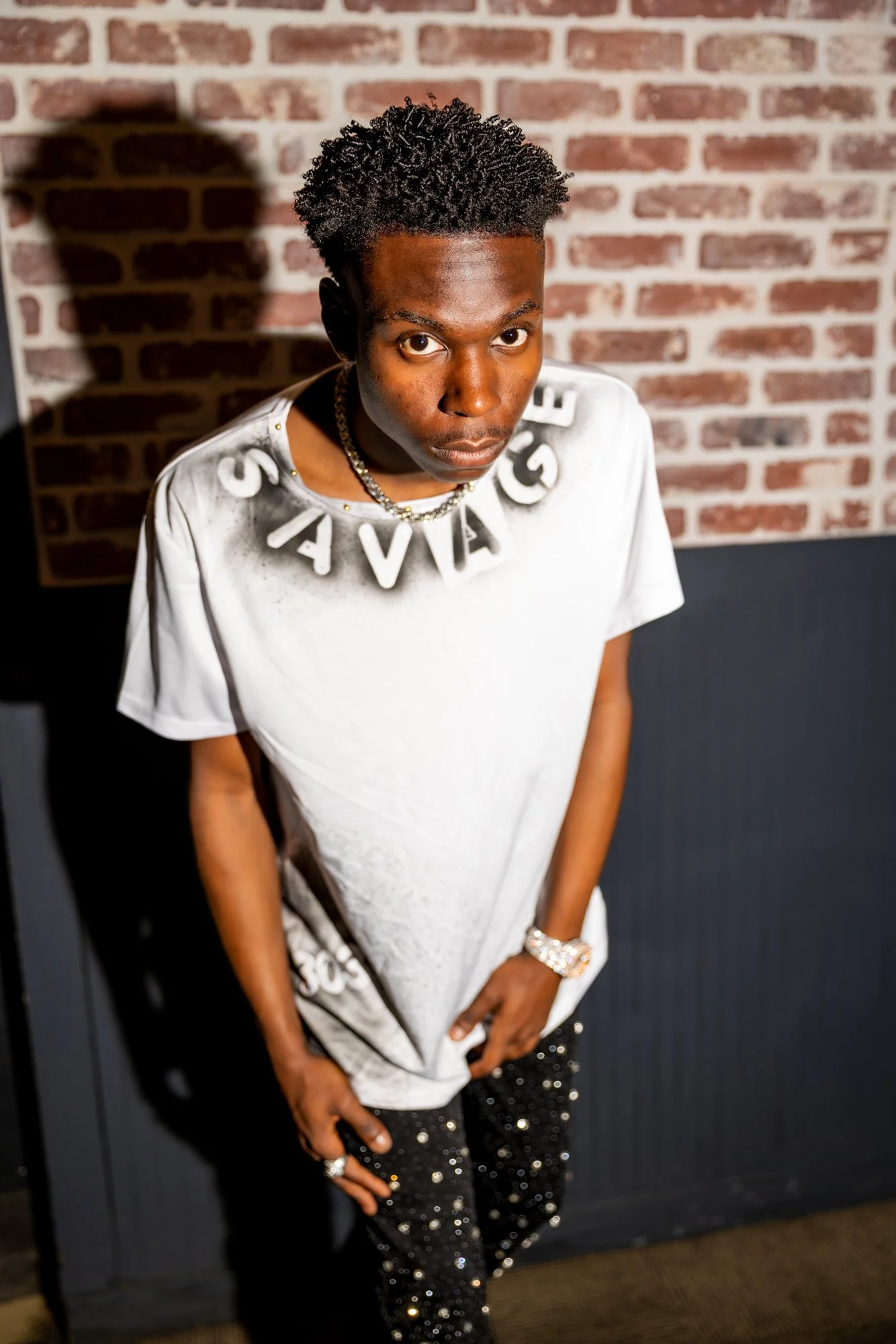 Young man with styled curly hair wearing a white T-shirt with large letter necklace and sparkling black pants, standing against a brick wall background.