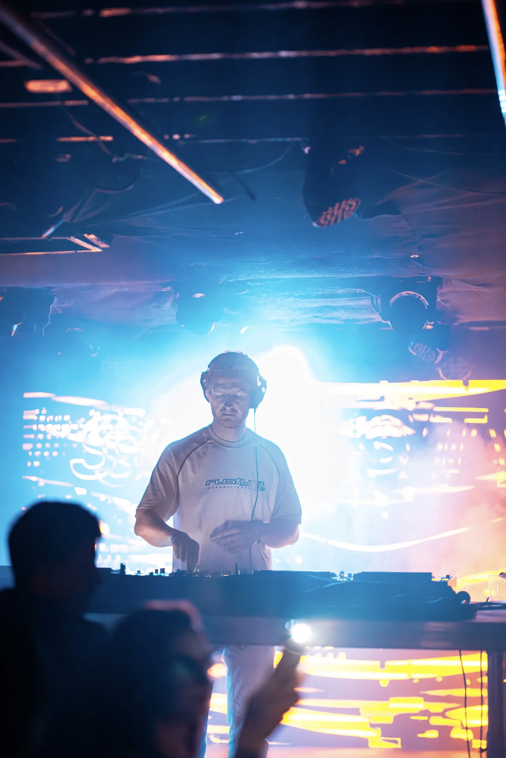 A DJ wearing a white Rush International shirt stands behind a DJ mixer on a brightly lit stage with colorful LED lights and visual graphics in the background.