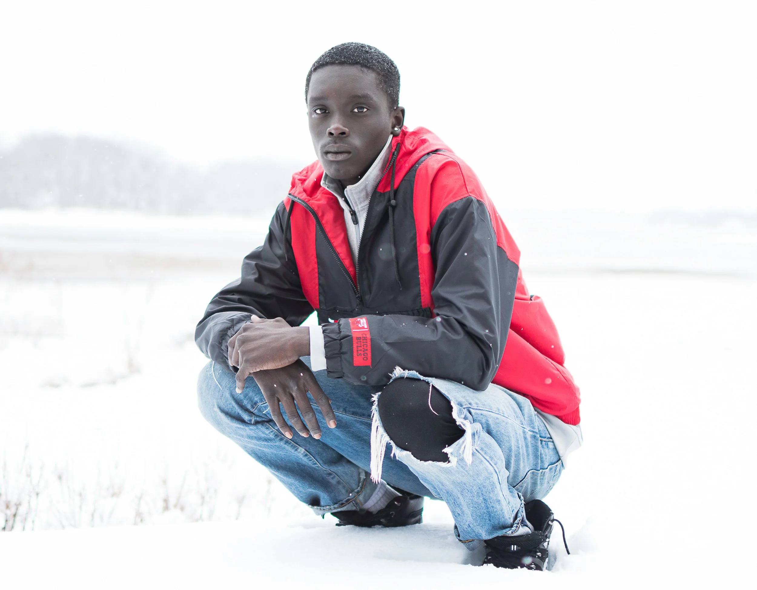 A young man with dark skin crouching in the snow outdoors, wearing a red and black jacket, ripped jeans, and black boots, with mountains and snow in the background.