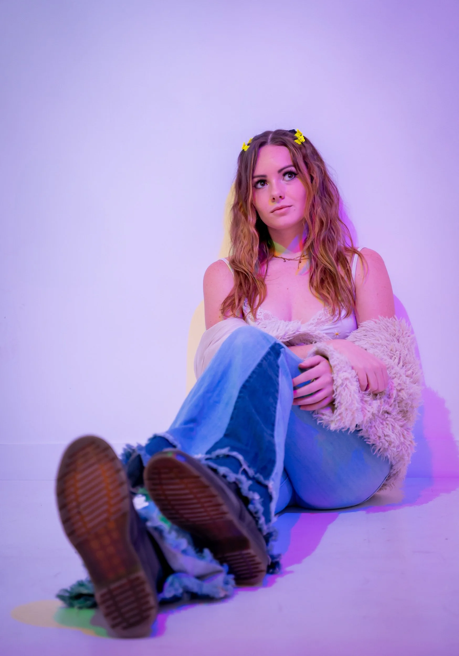A young woman with reddish-brown hair styled in loose waves, wearing a yellow butterfly hair clip, a white lace top, and a fluffy beige jacket, sitting on the floor with her legs crossed, looking thoughtfully to the side against a plain light purple 