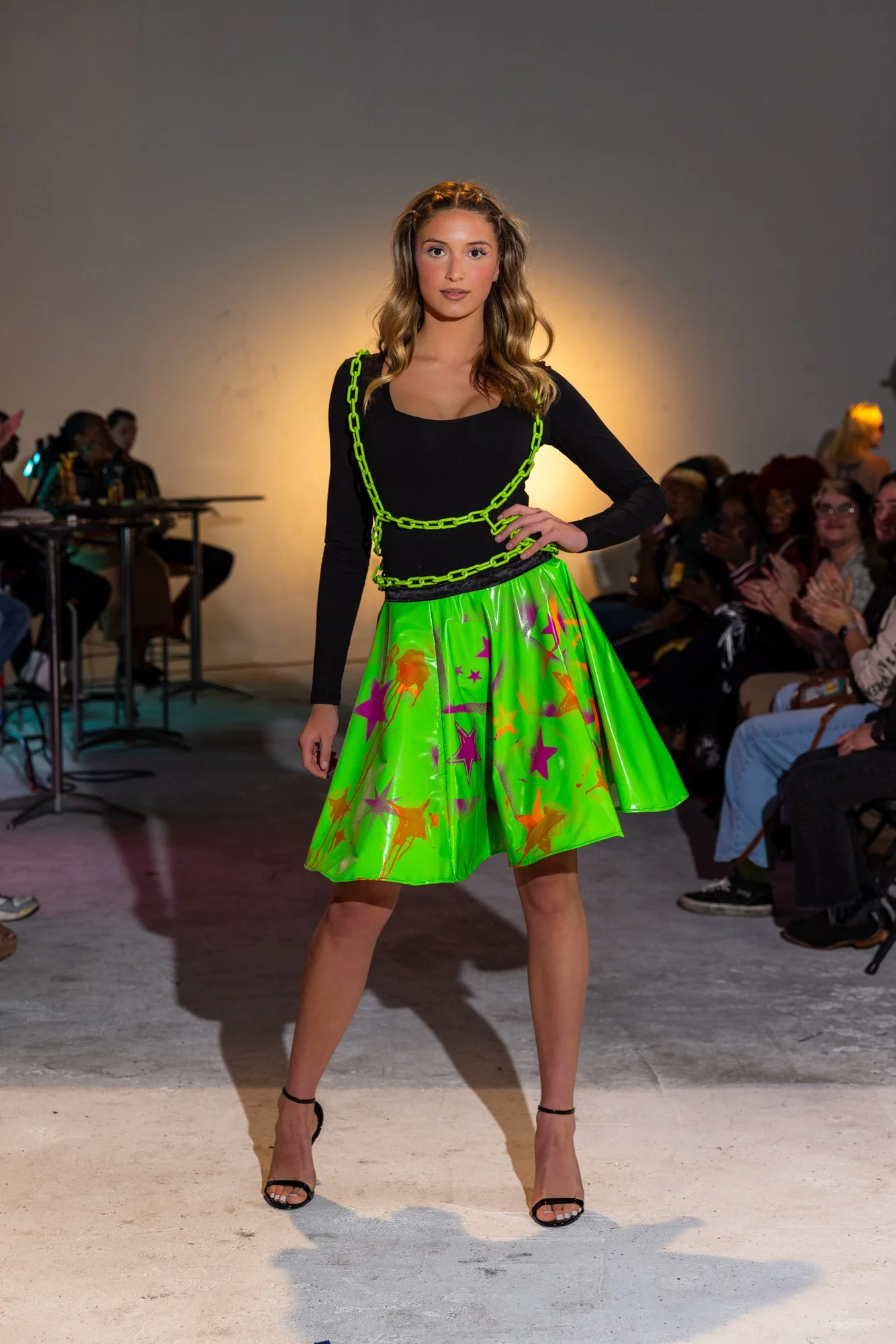 A fashion model walking down the runway in a colorful skirt with stars, paired with a black top, accessorized with a bright green chain necklace, and wearing black high heel shoes. The audience is watching and clapping in the background.