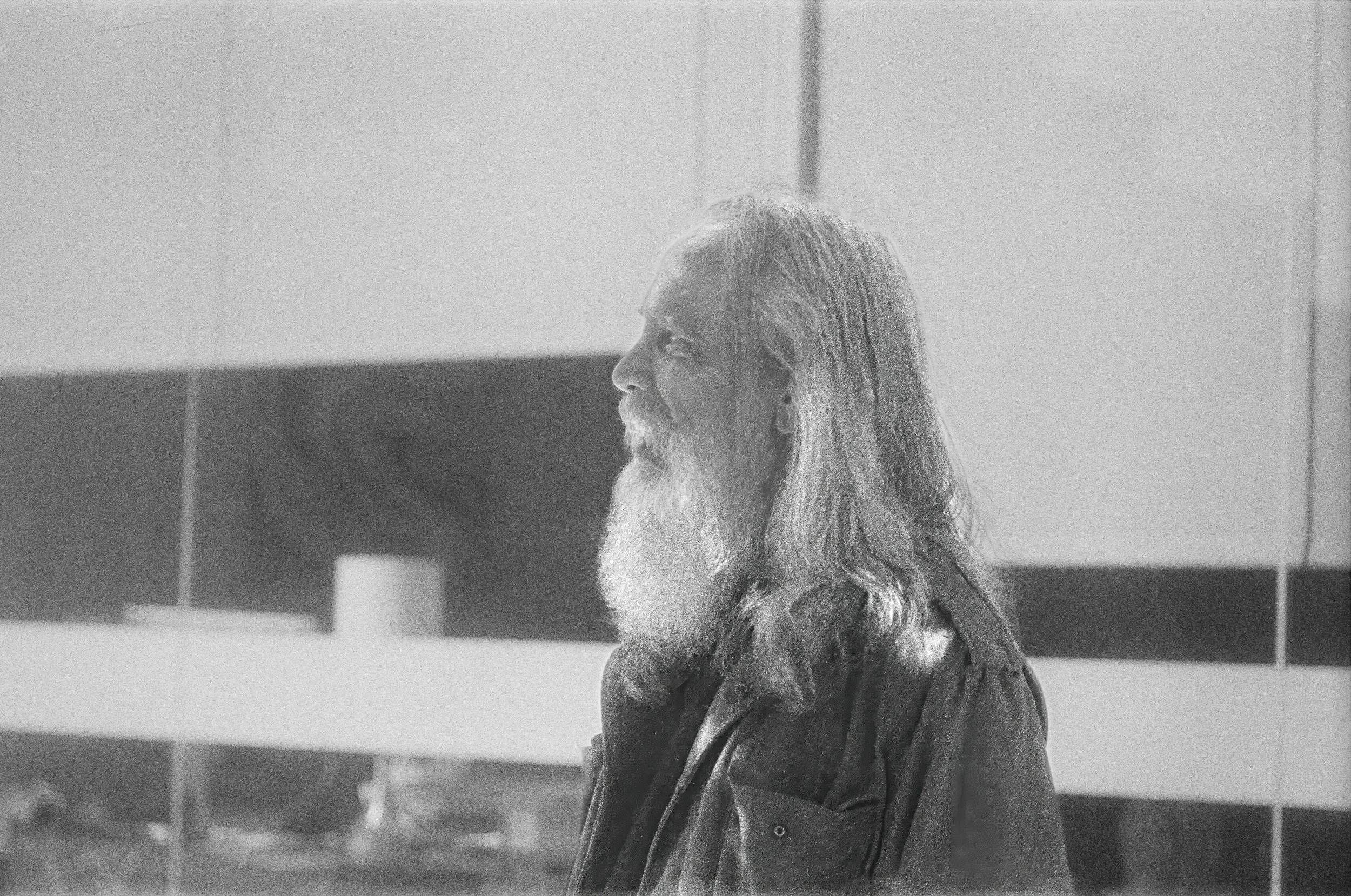 A black-and-white photo of a man with long hair and a beard, possibly in a bathroom or kitchen, with tiled walls in the background.