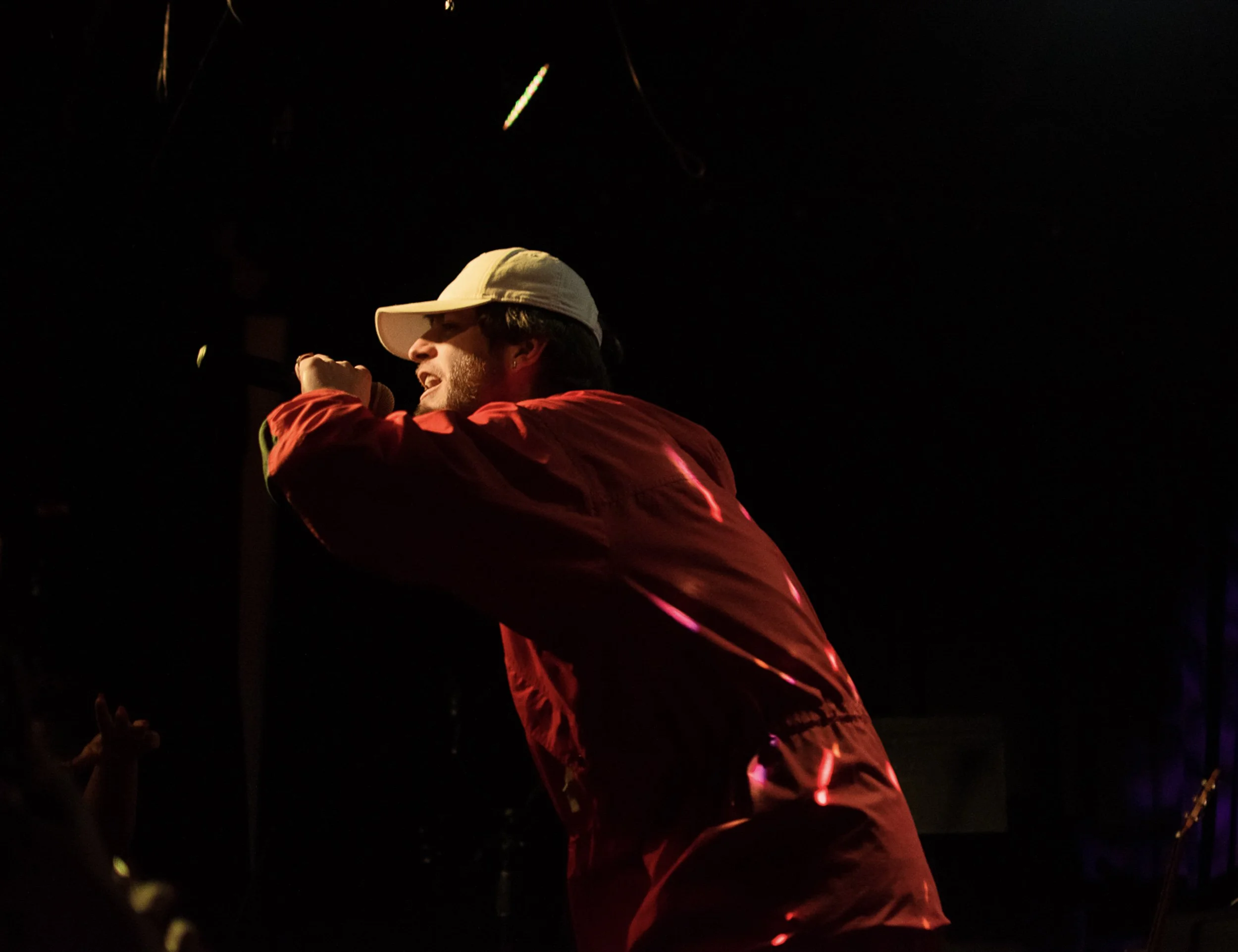 A person wearing a beige cap and a red jacket, holding a microphone and possibly singing or speaking, against a dark background with faint light reflections.