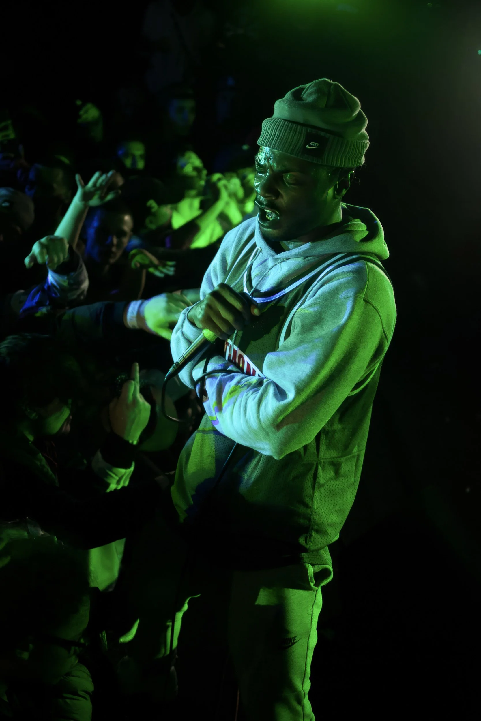 A man wearing glasses, a gray hoodie with a Nike logo, gray sweatpants, a green beanie, and holding a microphone, surrounded by a crowd at a concert or event with green lighting.