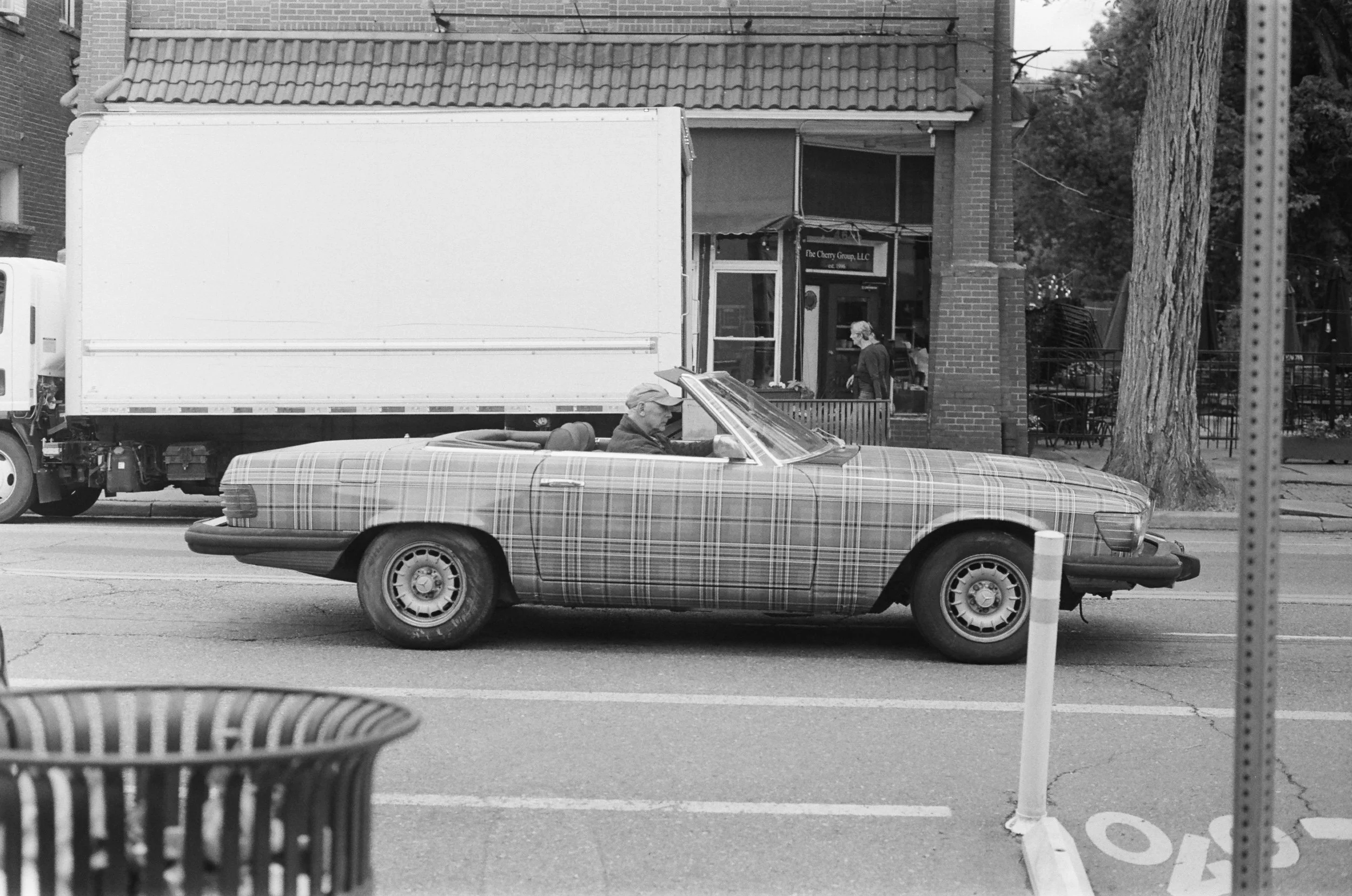 Black and white street scene with a vintage car in the foreground and a truck passing by. A person is visible inside the car, and another person is walking near a building with brick walls and a tiled roof.