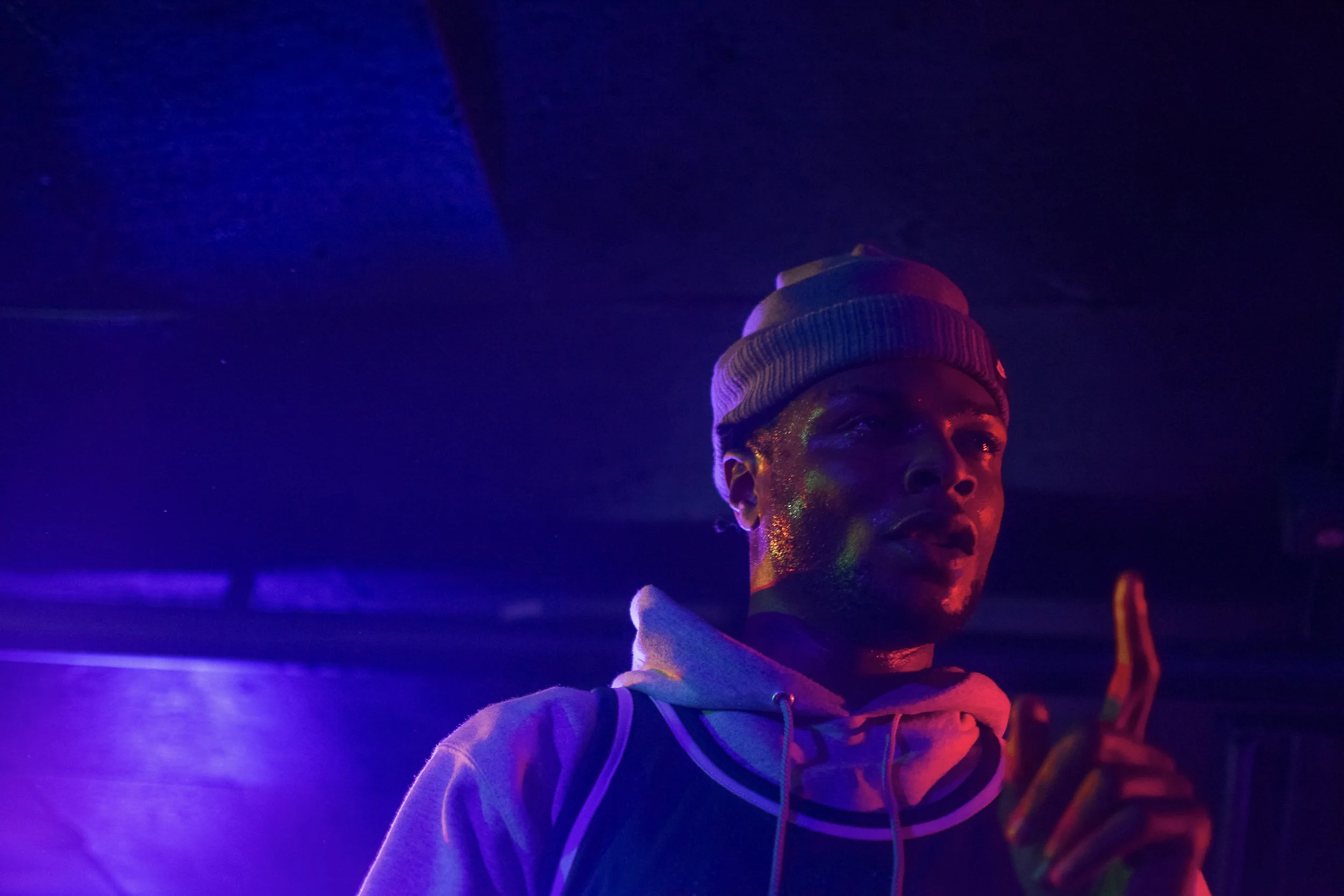 Portrait of a woman with colorful face paint wearing a beanie and hoodie, illuminated by purple and blue lighting.