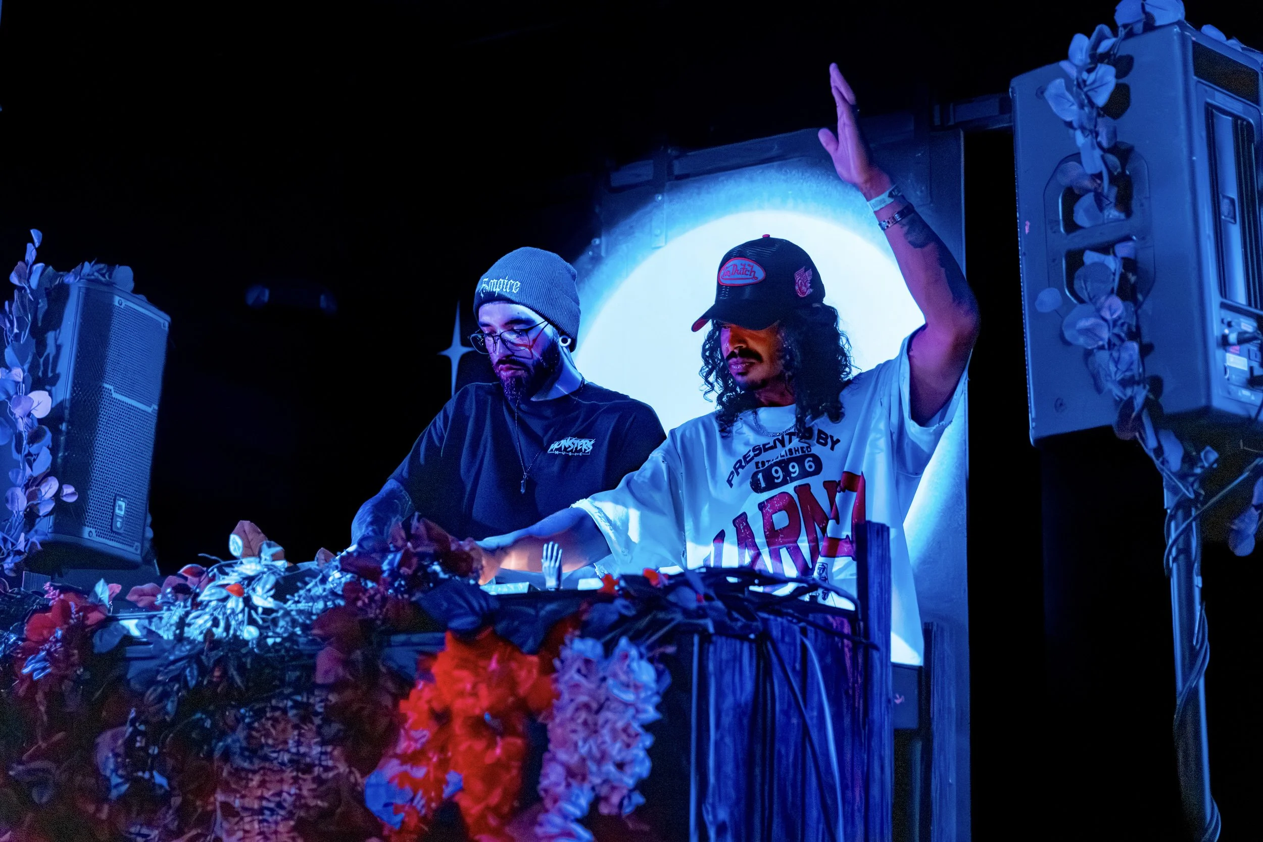 Two DJs performing at a nighttime event with blue lighting and floral decorations.
