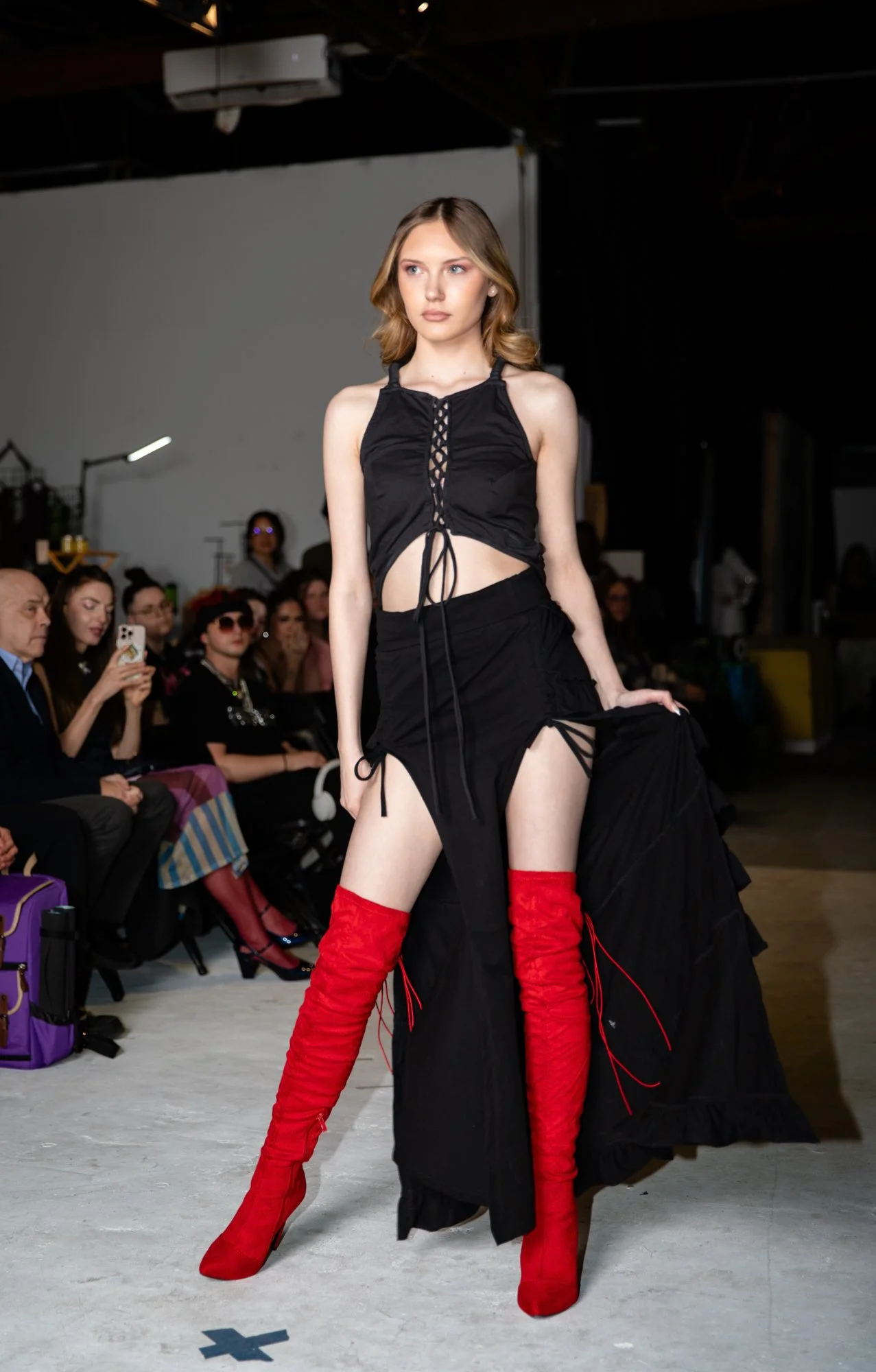 A female model walking on a runway during a fashion show, wearing a black top with lace-up details, a long black skirt with side slits, and red thigh-high boots. Audience members are seated along the runway, some taking photos with their phones.