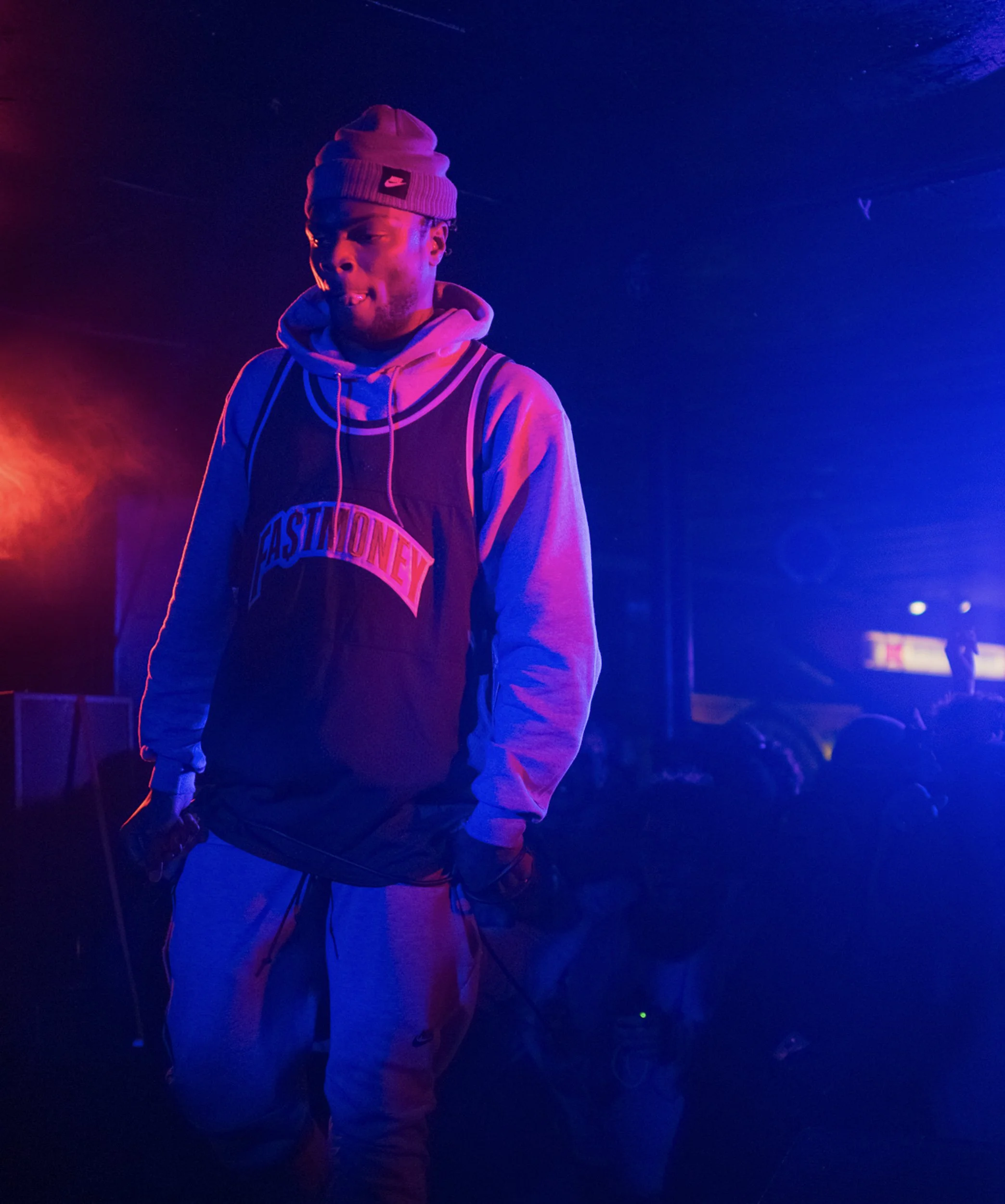 A young man wearing a Beanie, hoodie, and basketball jersey standing in colorful vibrant lighting, with a dark background.