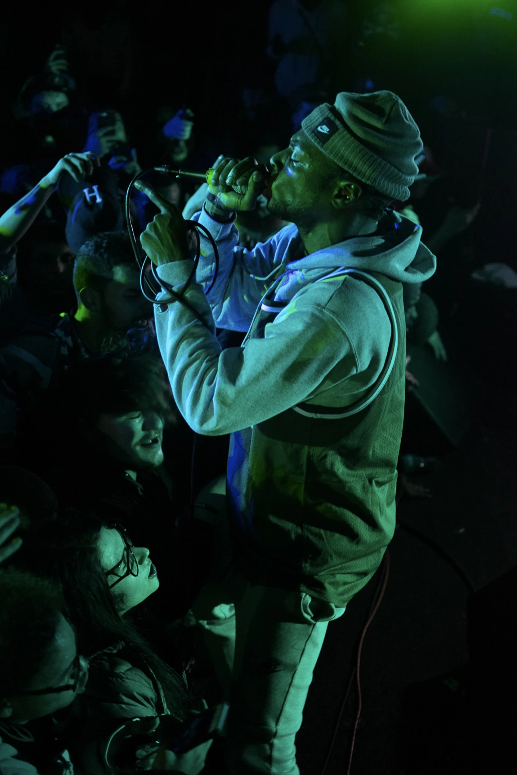 A man singing into a microphone at a concert, wearing a gray beanie and hoodie, with an audience watching and taking photos in a dark, colorful atmosphere.