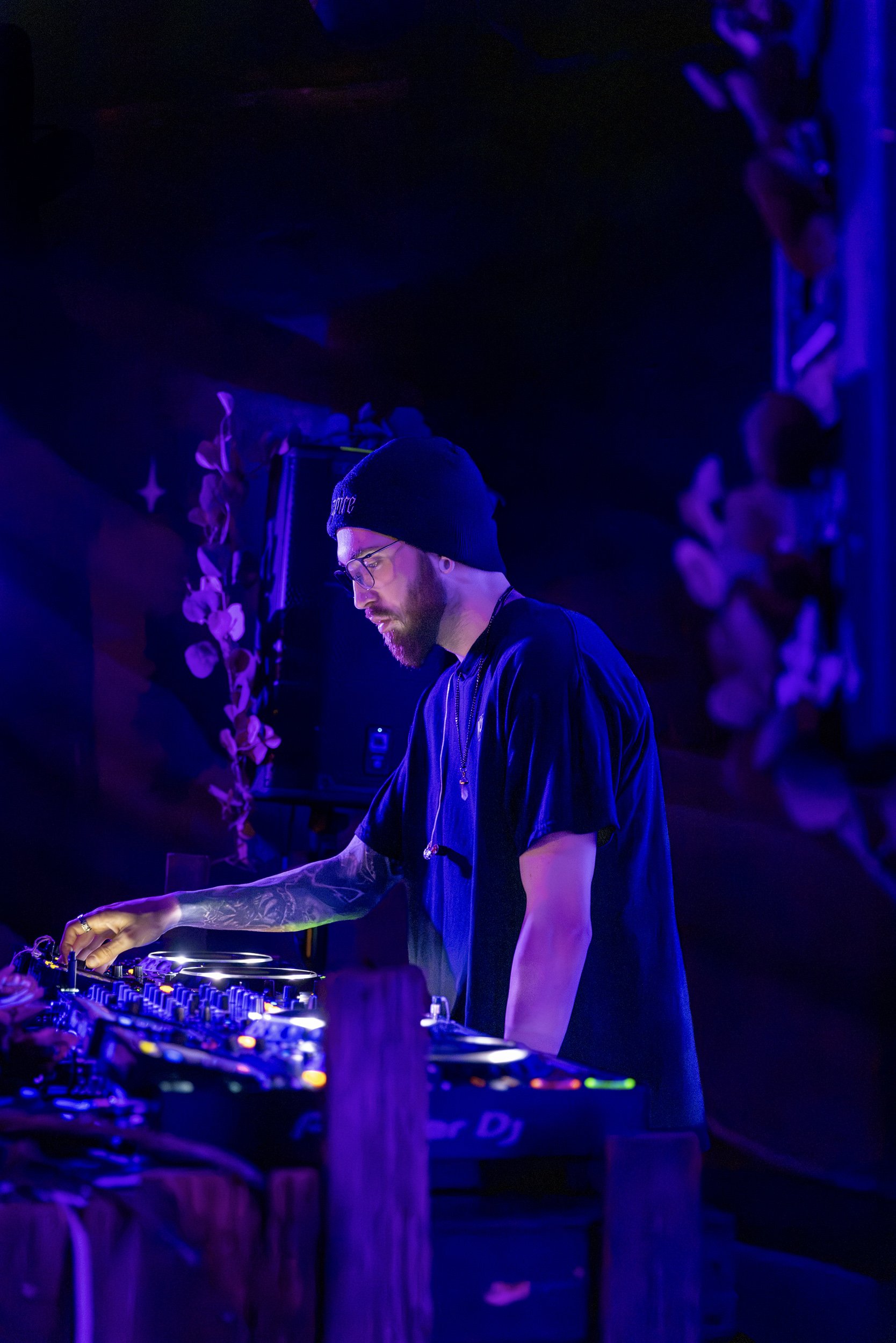 A DJ in a black beanie, glasses, and a black shirt is performing in a dark, purple-lit environment, adjusting equipment on a DJ console.