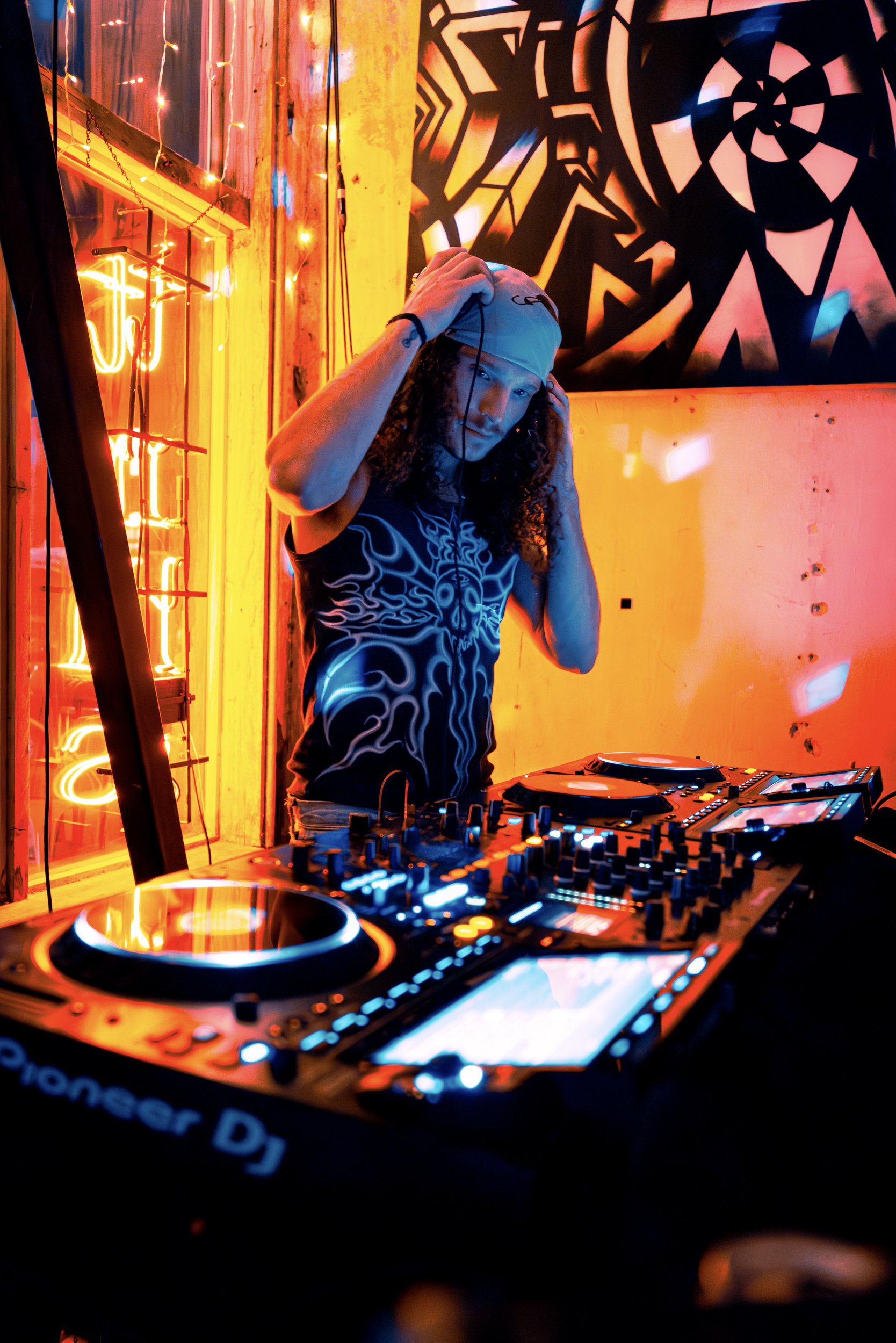 DJ with long curly hair wearing headphones and a black tank top with neon graphic, adjusting headphones in a vibrant, colorful nightclub setting with neon lights and geometric wall art.