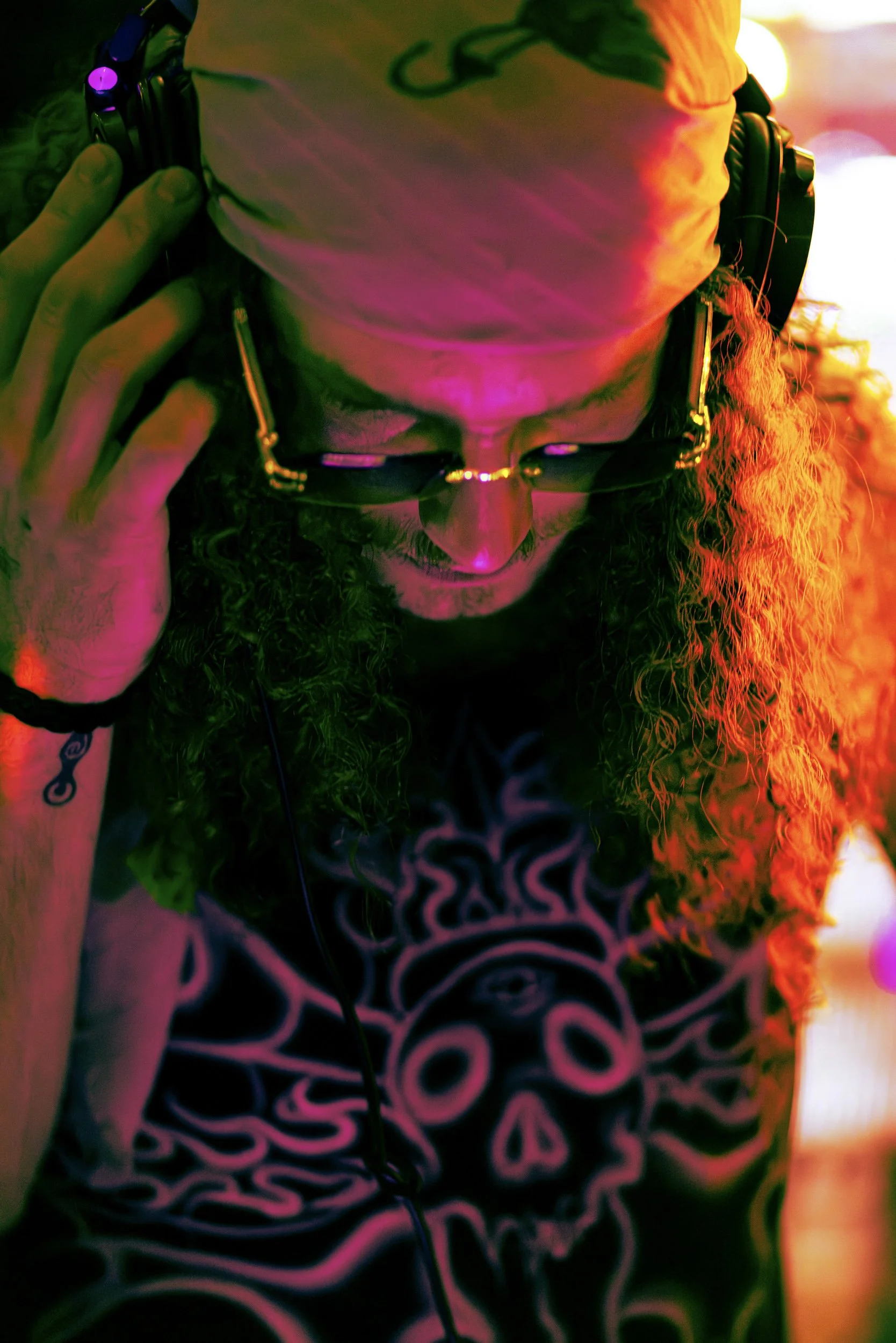 A person wearing glasses and a hoodie, with long curly hair. They are holding headphones to their ears and are illuminated by colorful lighting.