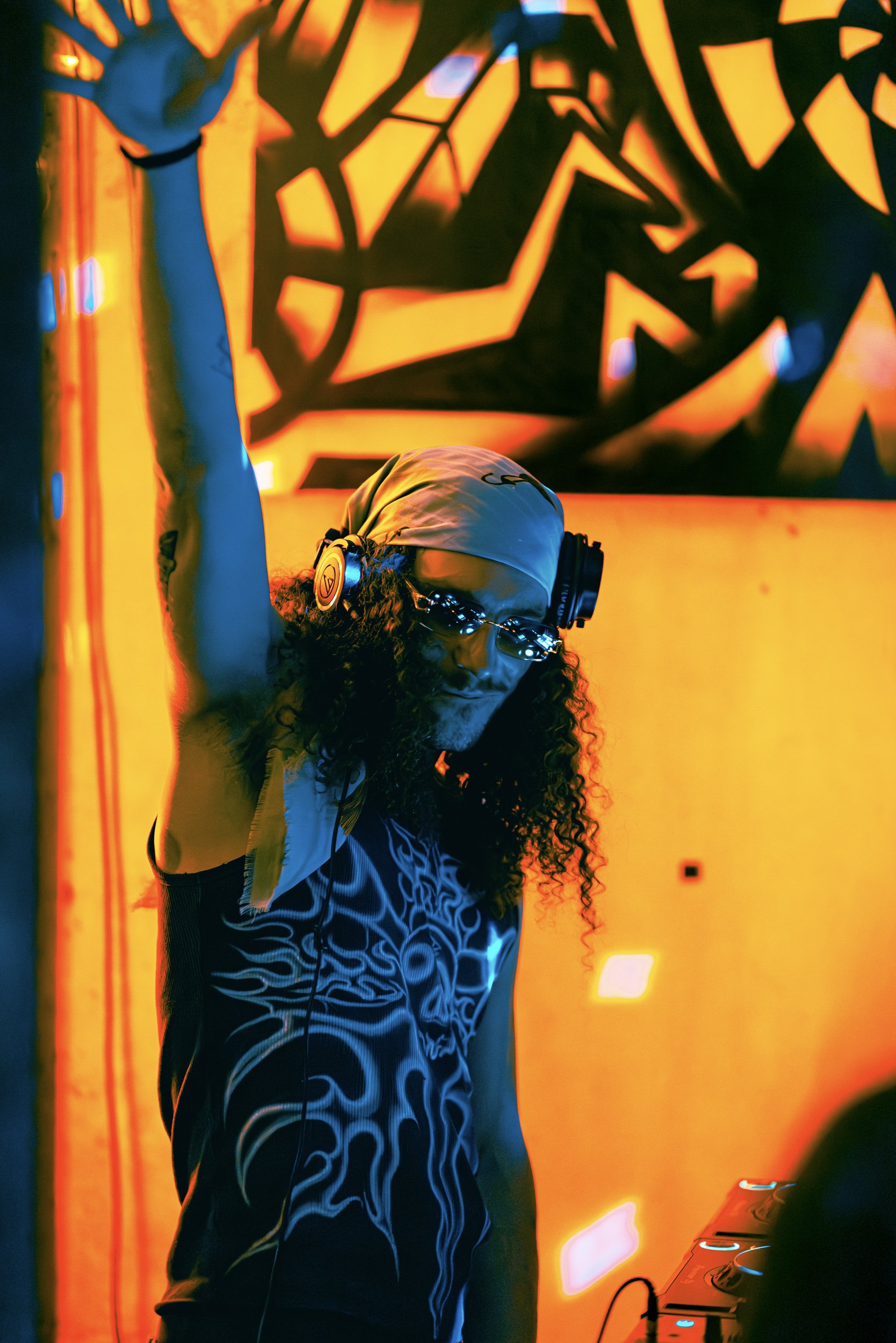 A DJ wearing headphones and sunglasses performing in a vibrant, colorful environment with abstract, bright orange and yellow lighting patterns.