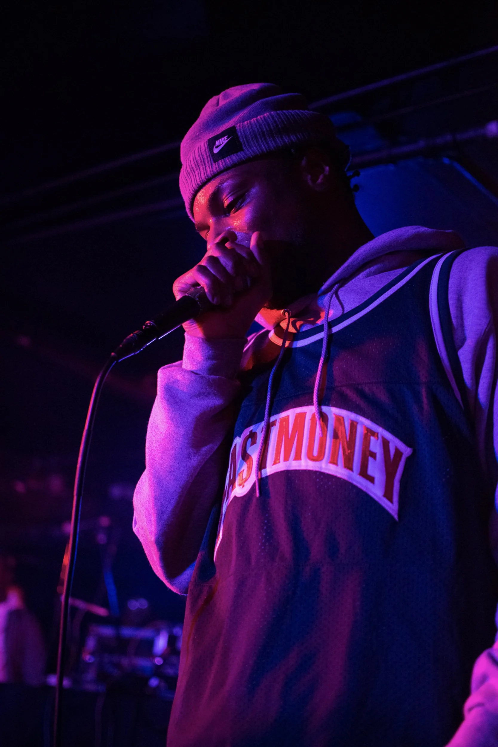 A young man wearing a gray Nike beanie and a hoodie with a 'STONEY' logo, holding a microphone, performing on stage illuminated by purple and pink stage lights.