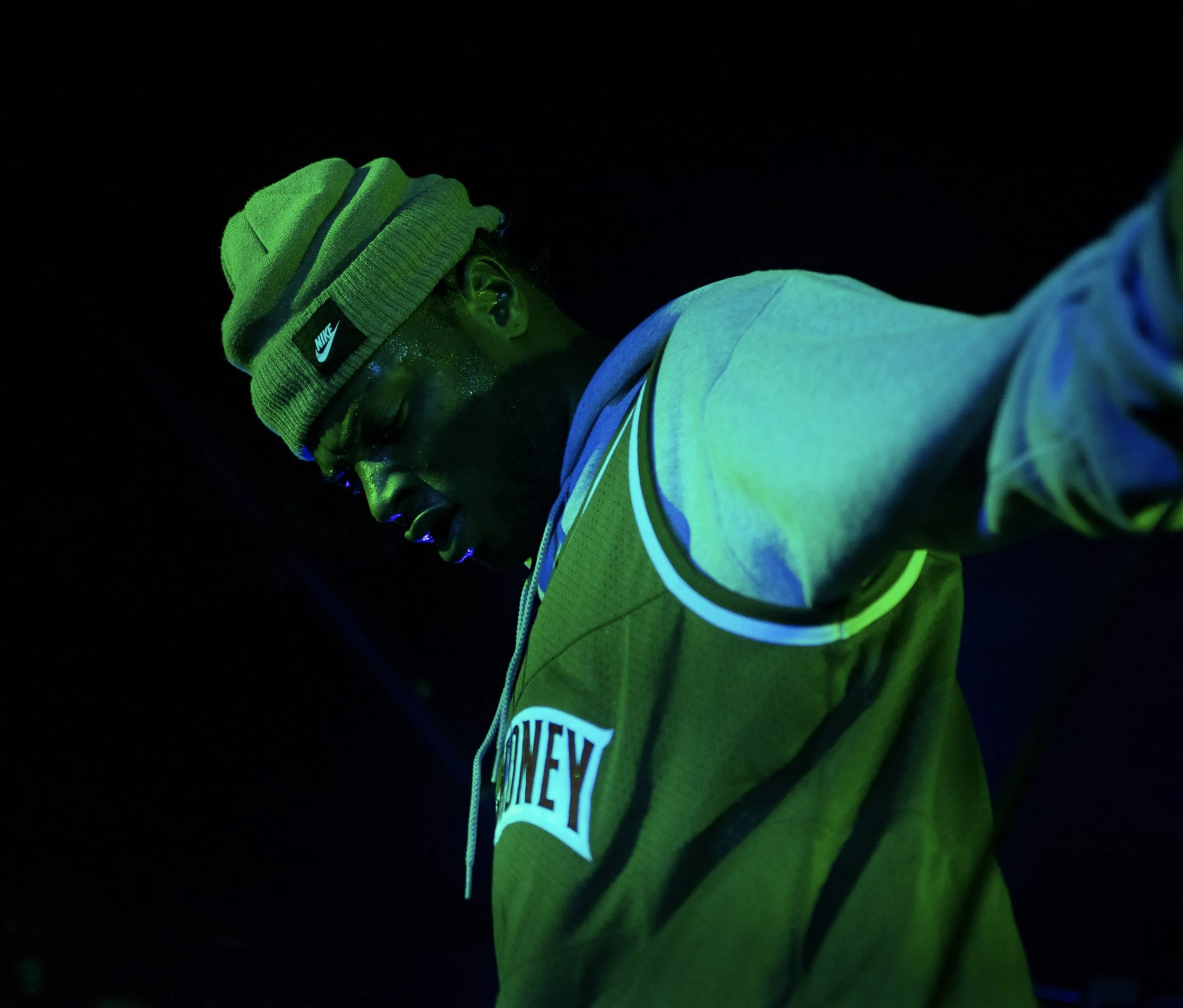 Person wearing a neon green Nike beanie and a yellowed tank top, illuminated with green and blue UV light.