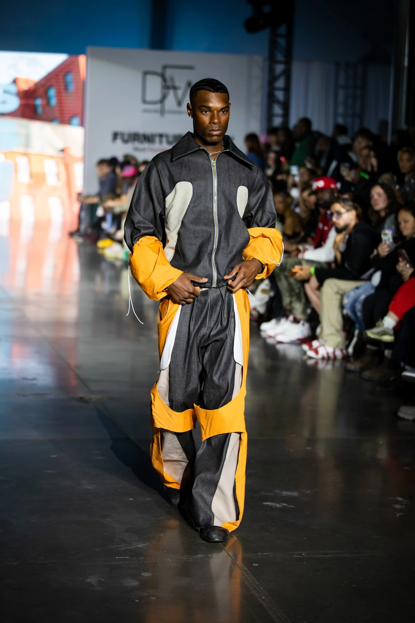 A male model walking down a runway at a fashion show, wearing a multi-colored jacket and matching pants, with an audience seated on either side of the runway.
