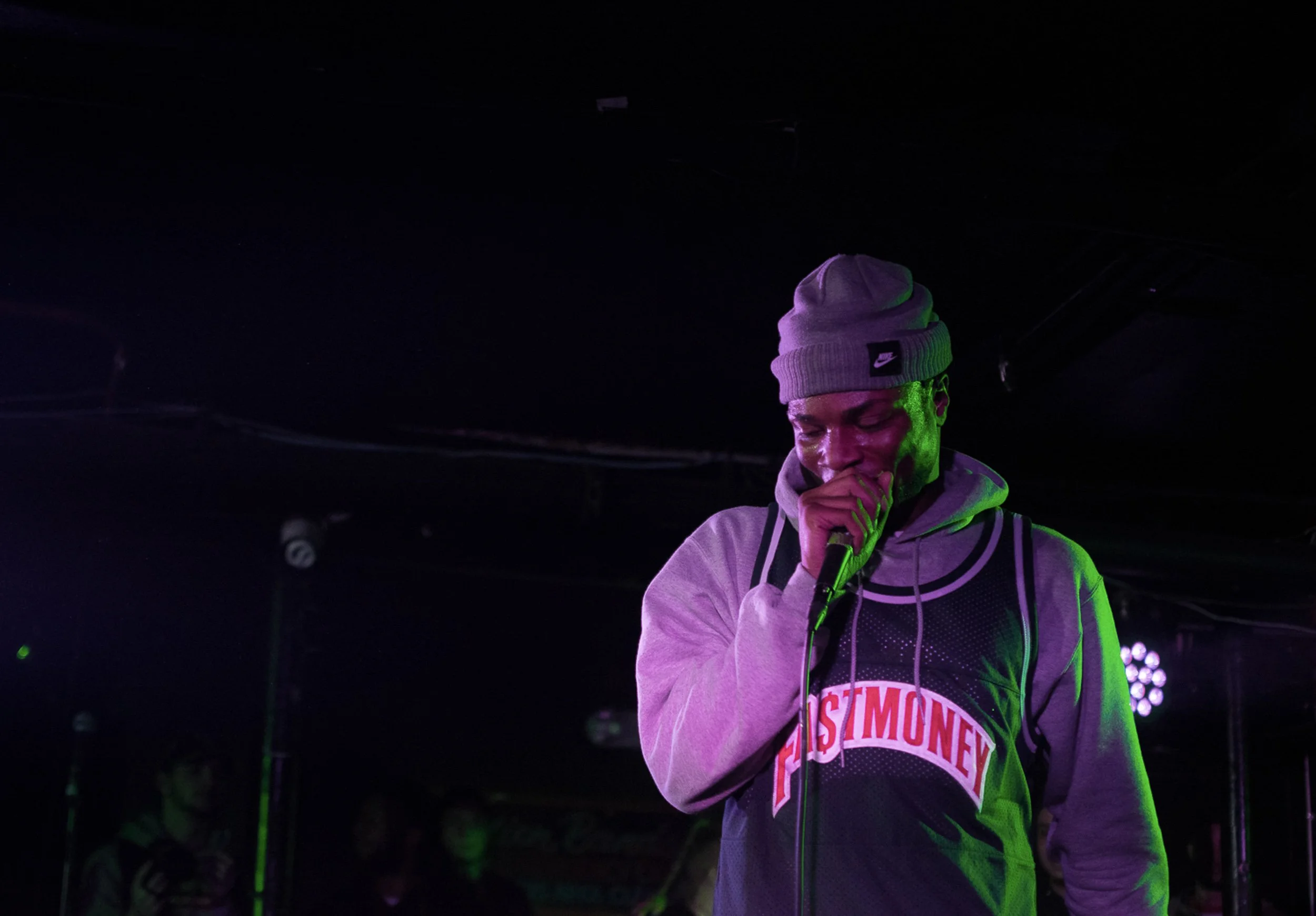 A young man wearing a grey beanie with a Nike logo and a hoodie with logo that reads 'FIST MONEY' stands on stage under purple and green lights, with his eyes closed and hand over his mouth.