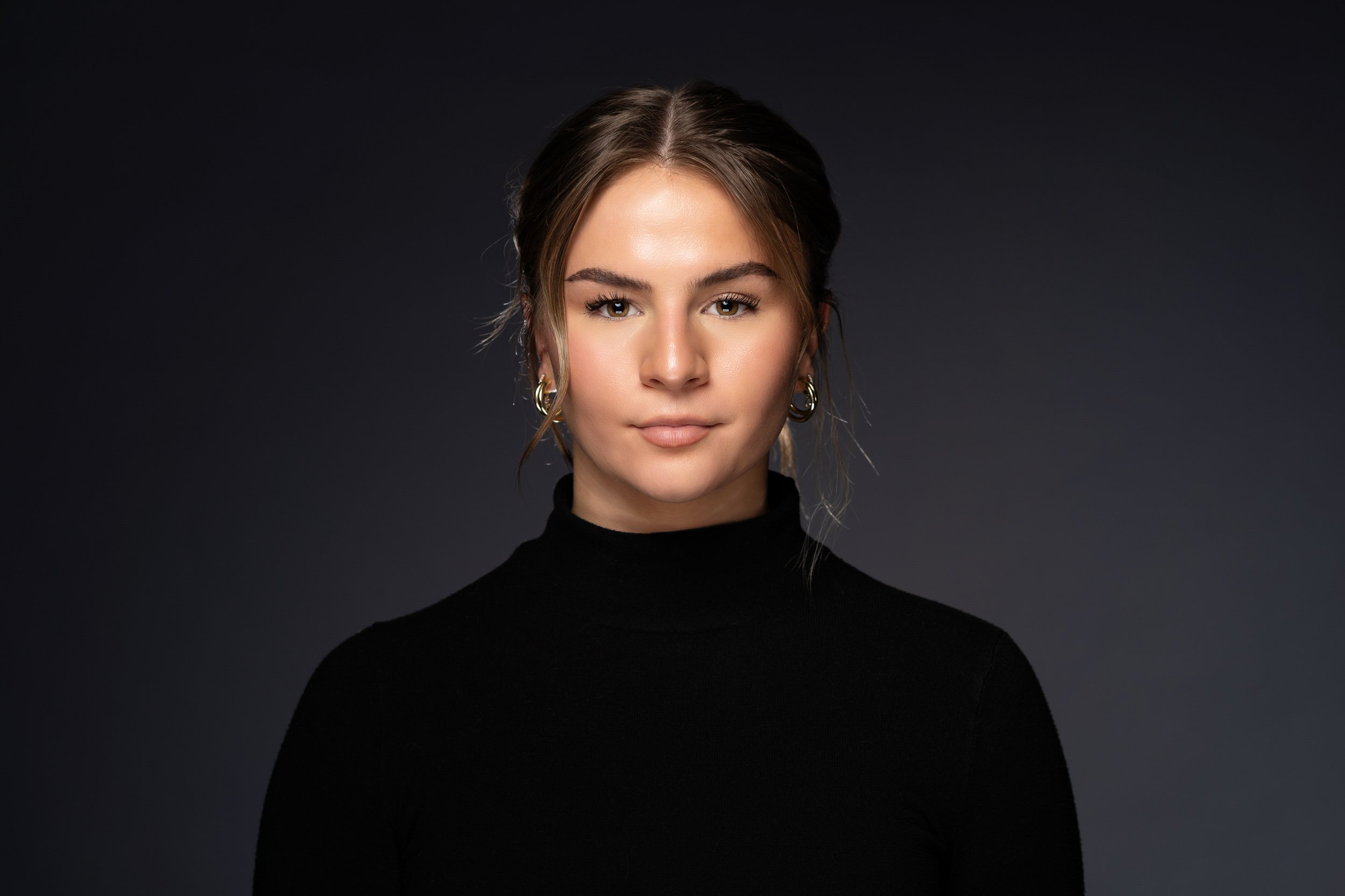 Portrait of a woman with brown hair styled in a loose updo, wearing a black turtleneck, gold hoop earrings, and neutral makeup, against a dark background.