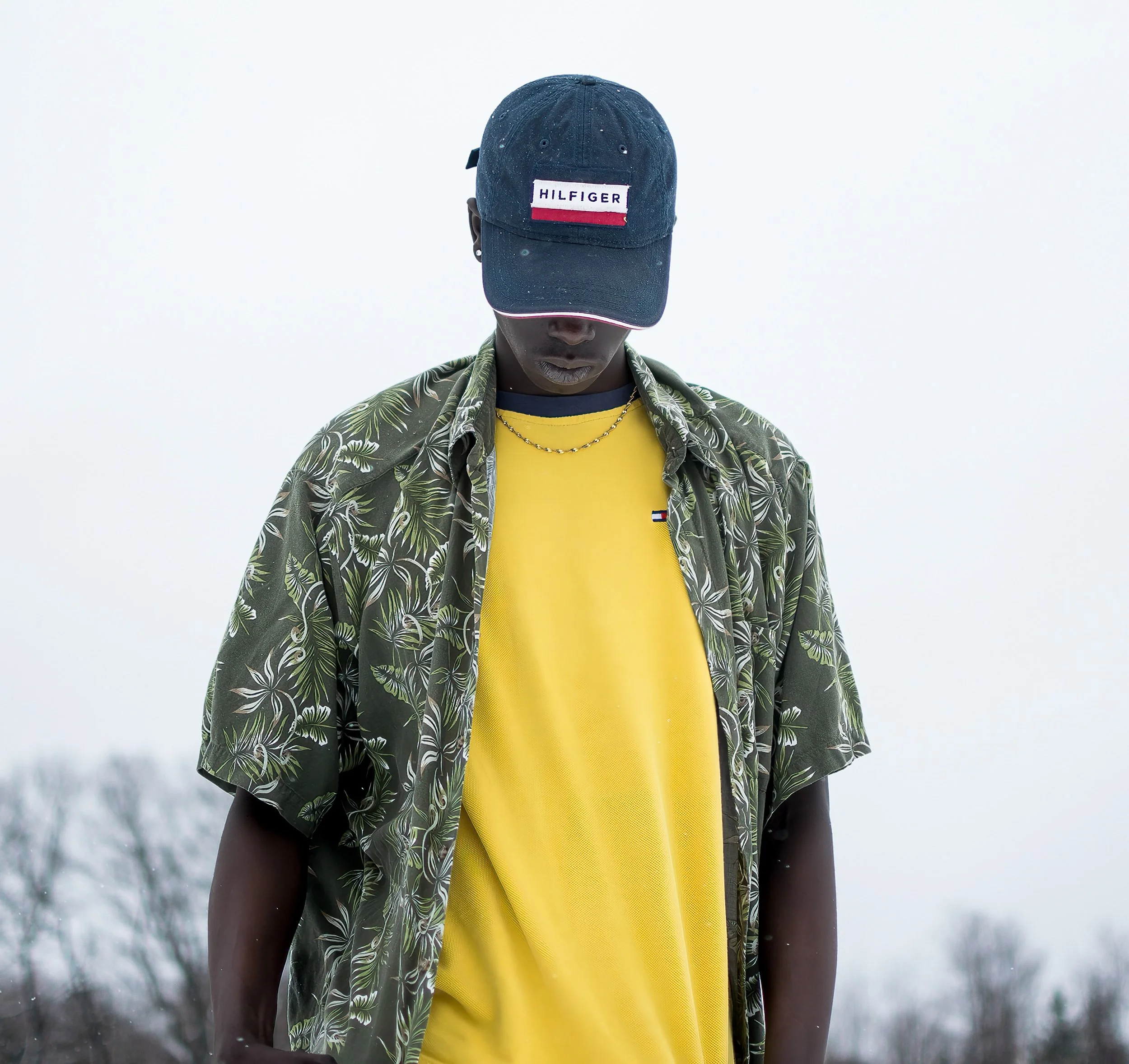 Person wearing a dark blue cap labeled 'Hilfiger' with rain droplets, yellow shirt, patterned green jacket, and gold chain, standing outdoors in snowy weather.