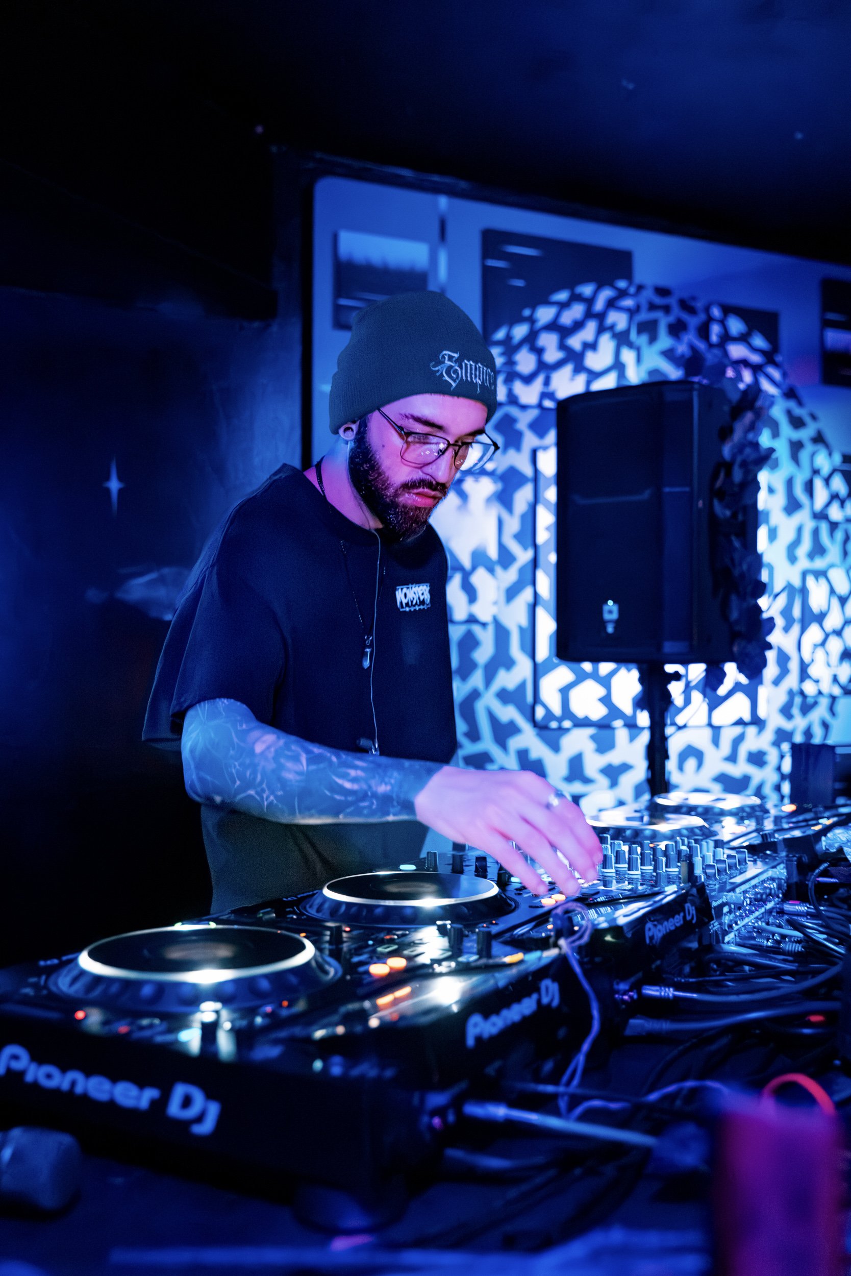 Male DJ with glasses and a green beanie playing music on DJ equipment in a club with blue and purple lighting.