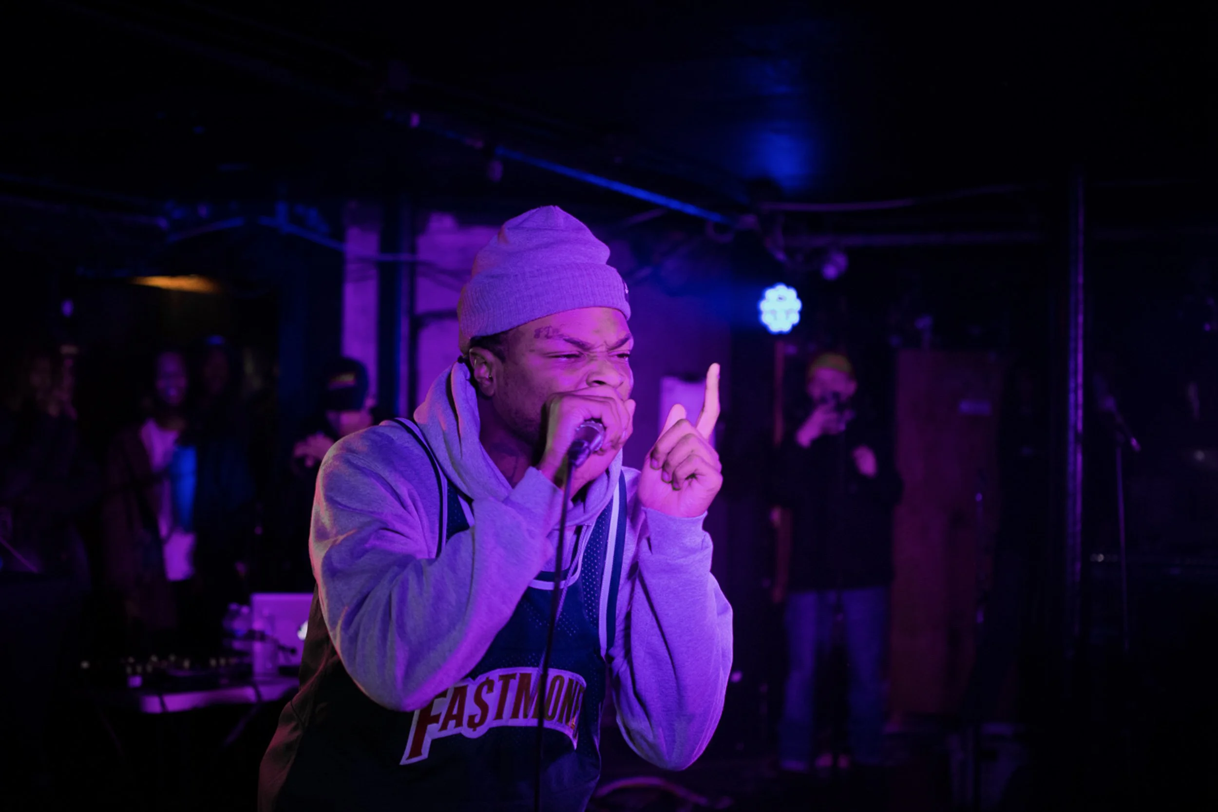 A young man wearing a gray hoodie and a gray beanie singing into a microphone on stage under purple and blue lights, with a blurred audience in the background.