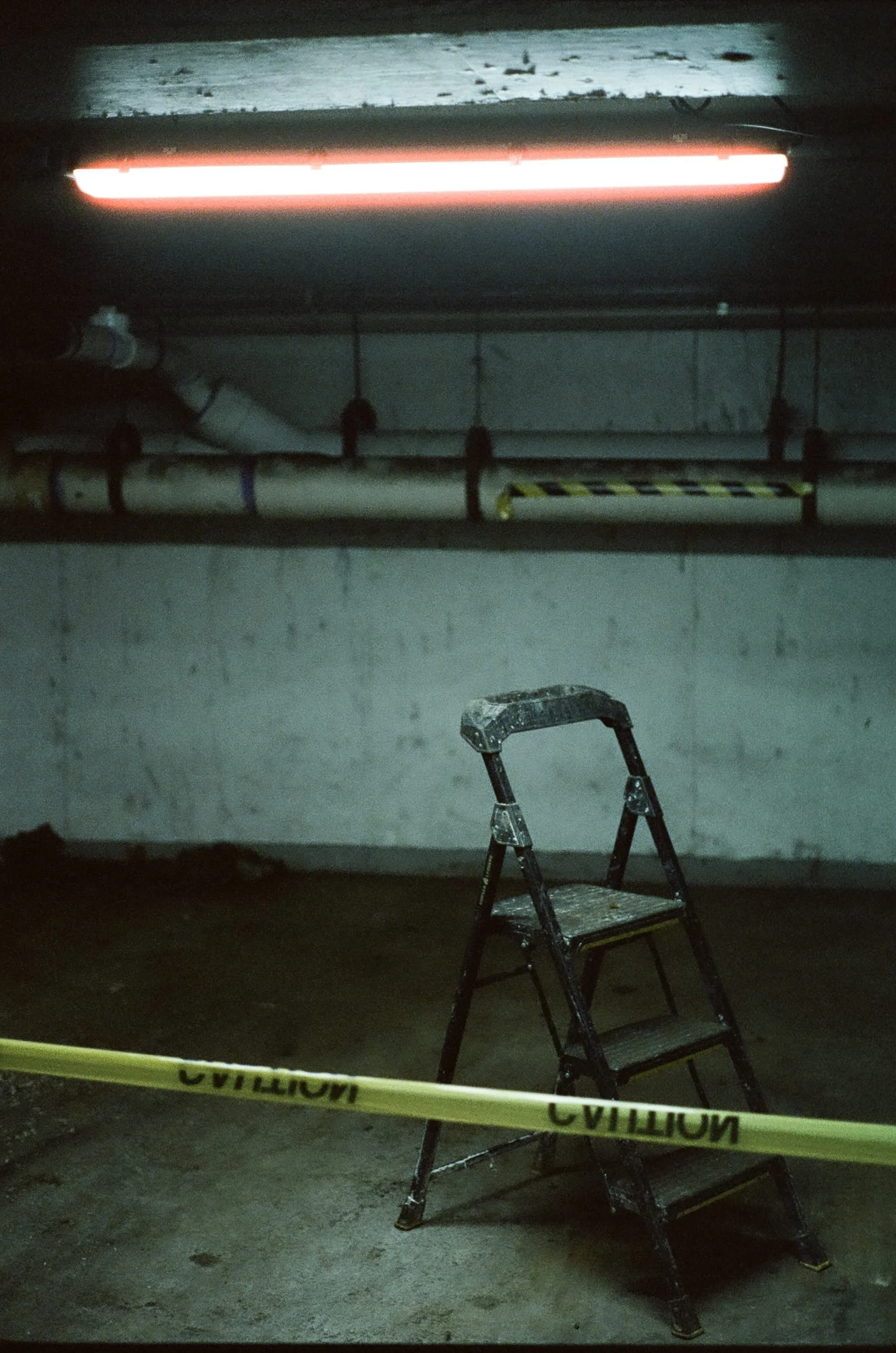 A dark underground parking garage with a red emergency light on the ceiling. In the foreground, a stepladder is positioned, surrounded by yellow caution tape on the floor. The garage has pipes running along the wall and some construction or maintenan